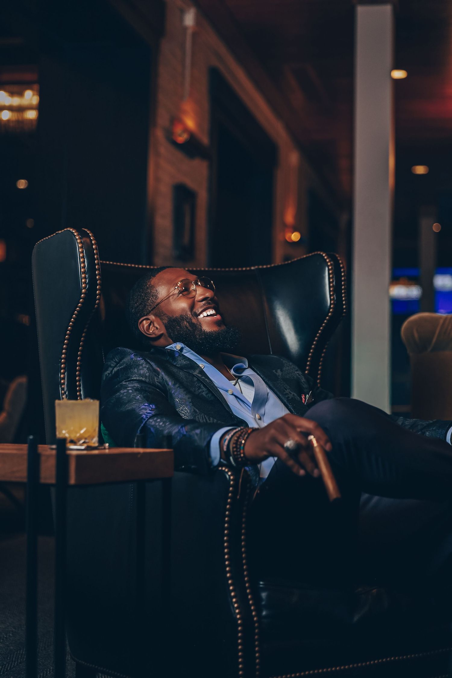 Stylish Black man in designer suit laughing joyfully while relaxing in leather chair holding cigar.