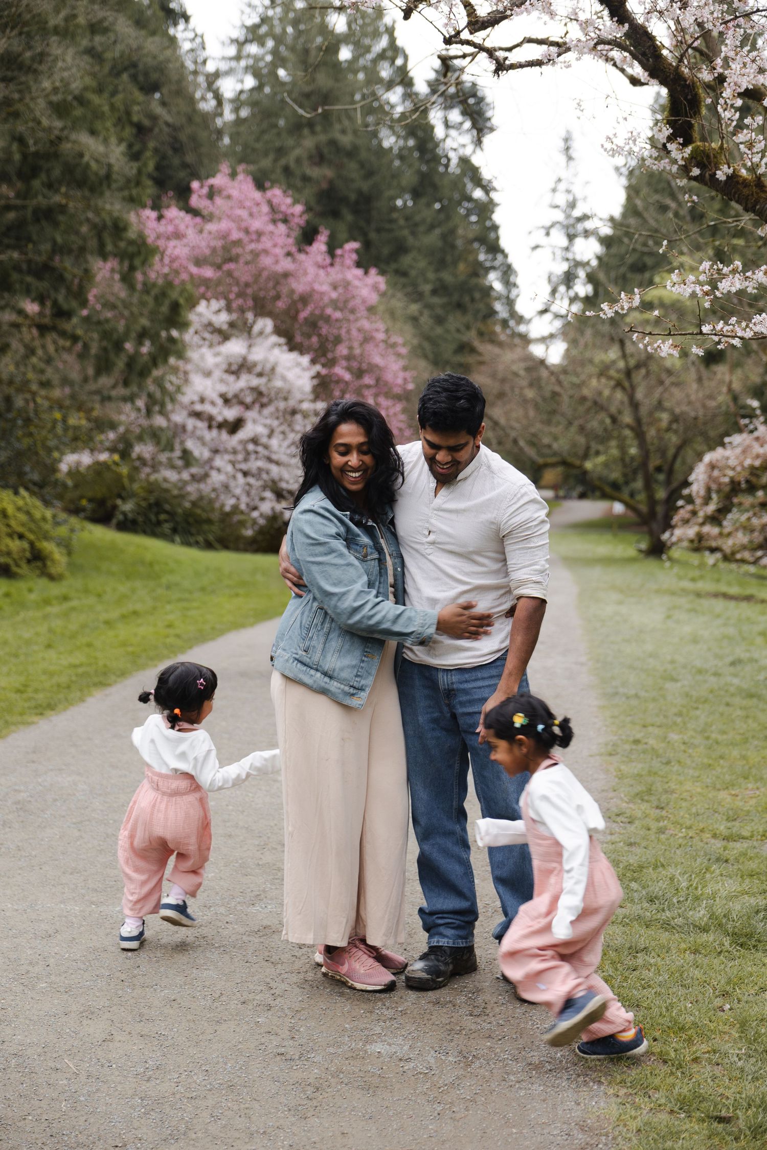 A joyful South Asian family with two toddlers embracing on a garden path surrounded by blooming cherry blossom trees.