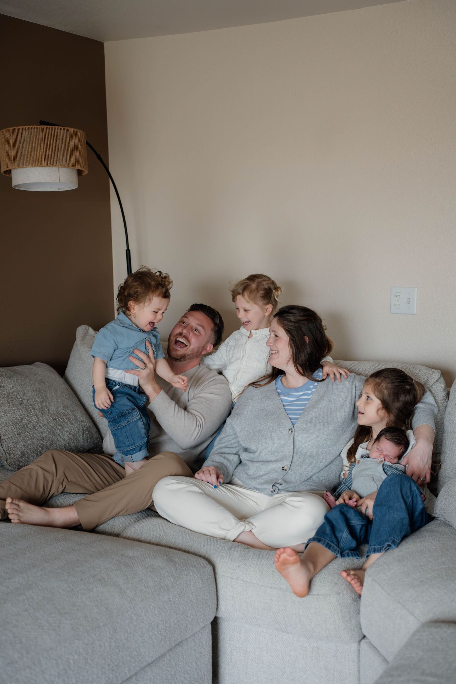 Happy family of five sitting on a gray couch, parents laughing with three young children in a cozy living room.