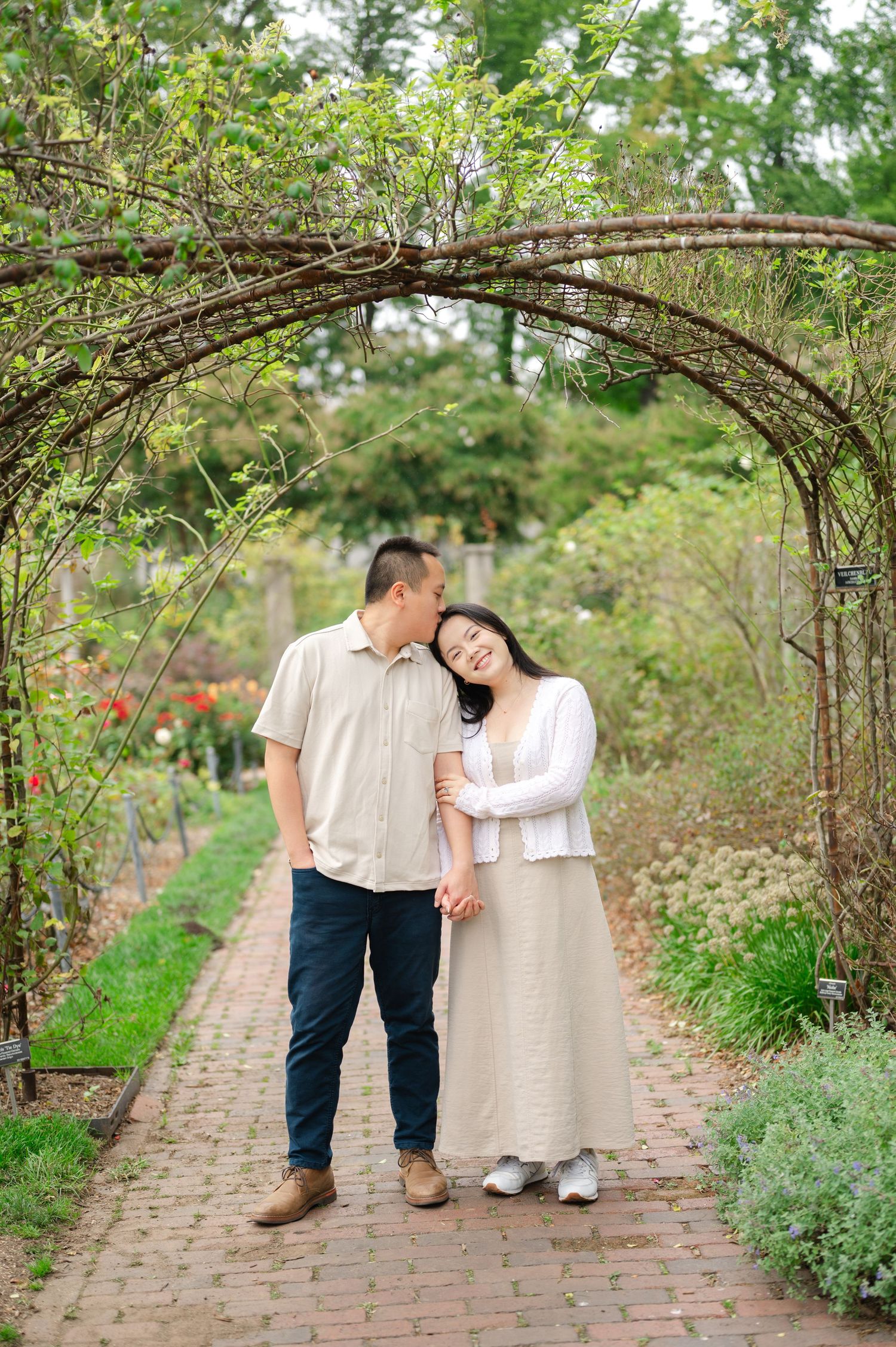 A man kisses his new fiancé on the head as they pose while walking through the rose garden of the Brooklyn Botanic Garden after a proposal.