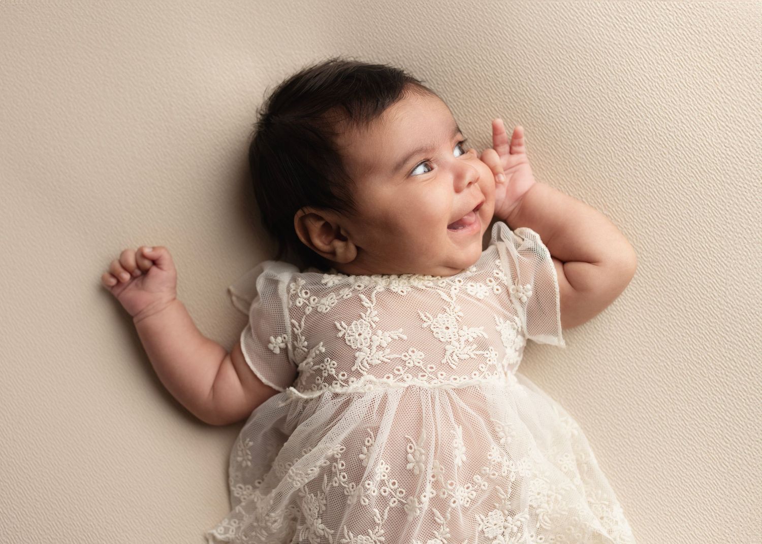 Adorable baby girl in white lace dress lying on cream background, looking up with curiosity.