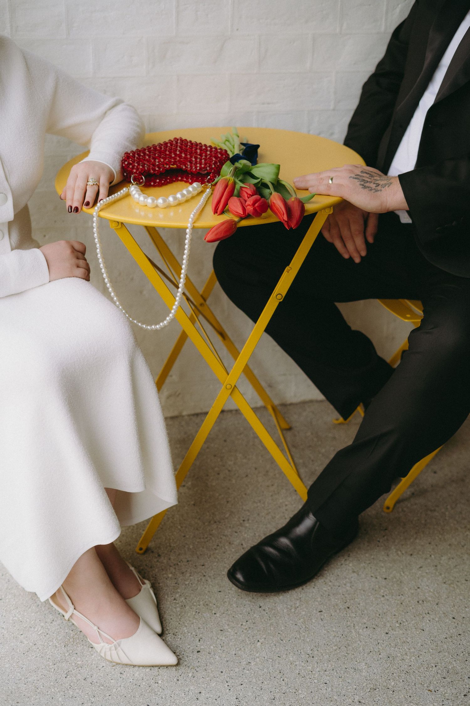 Bride and groom seated beside a yellow folding table with red roses and pearl necklace, wearing white dress and black suit.