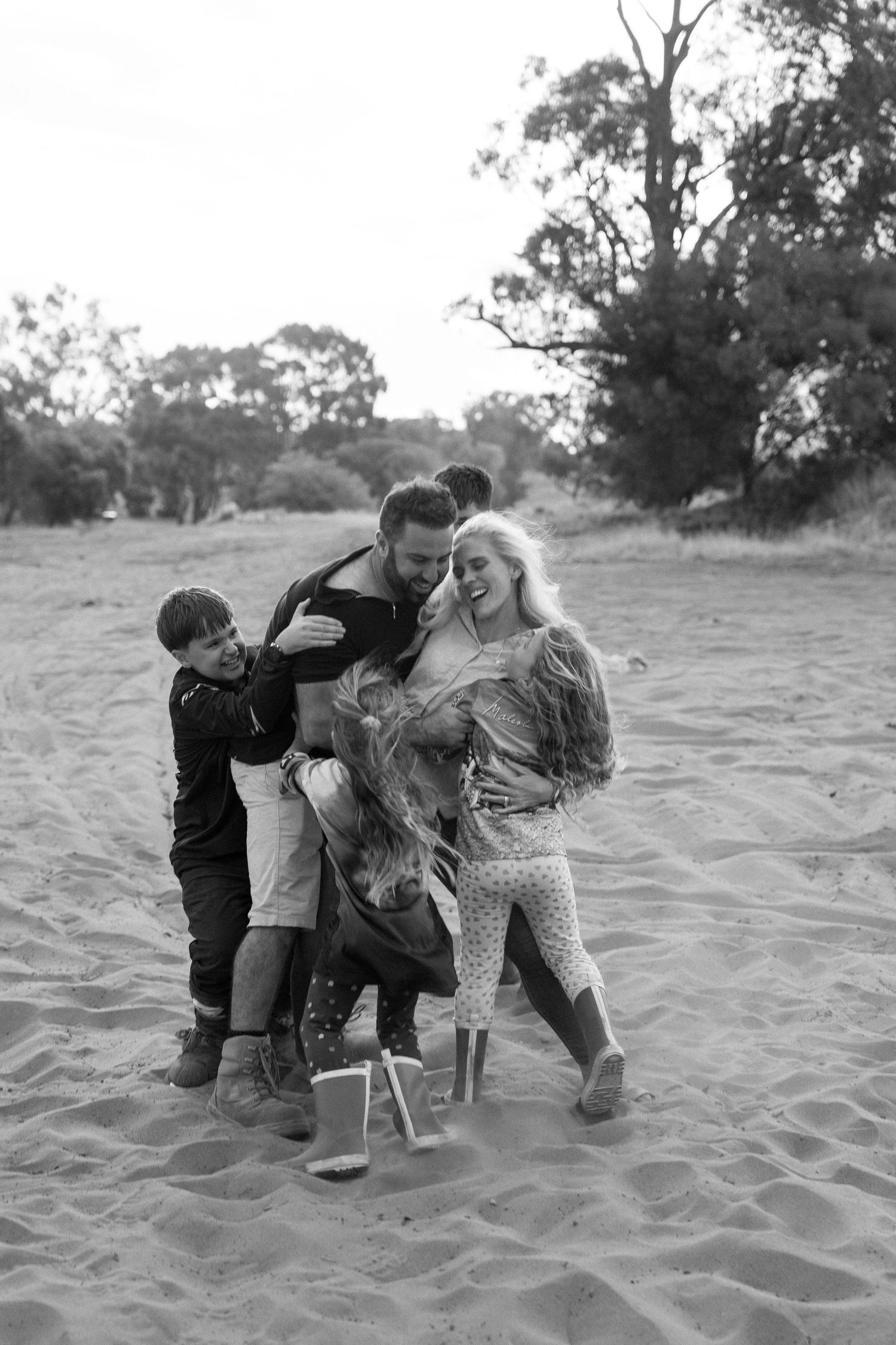 Black and white photo of a joyful family playing together on a sandy beach with trees in the background.
