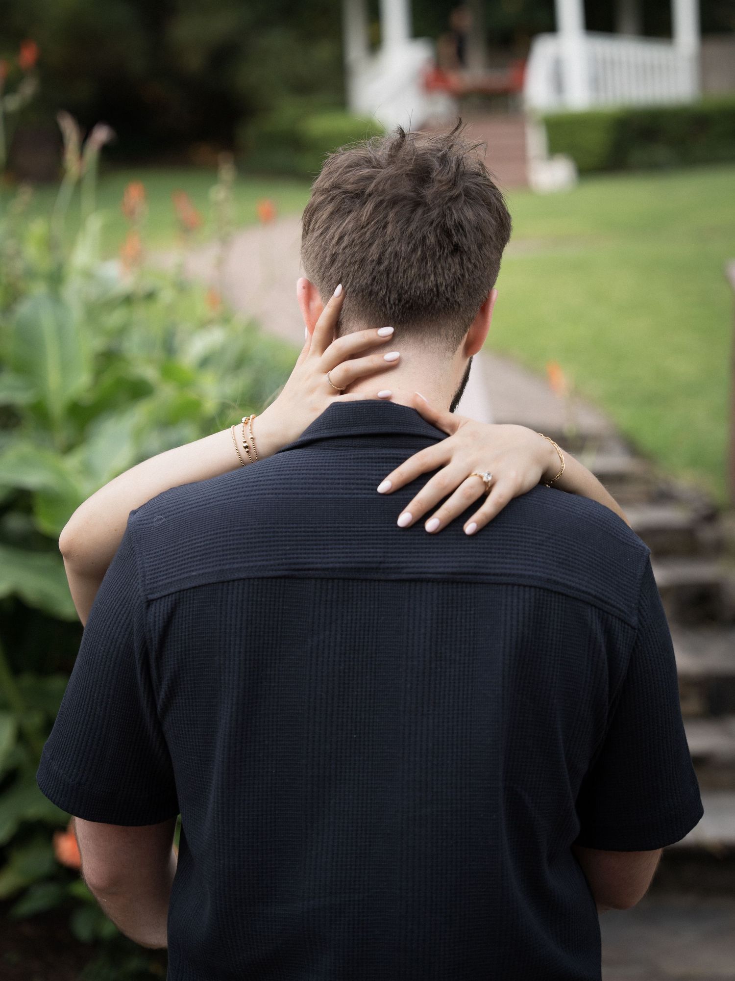 Couple embracing outdoors, woman's hand with gold bracelet and ring resting on man's shoulder in garden setting.