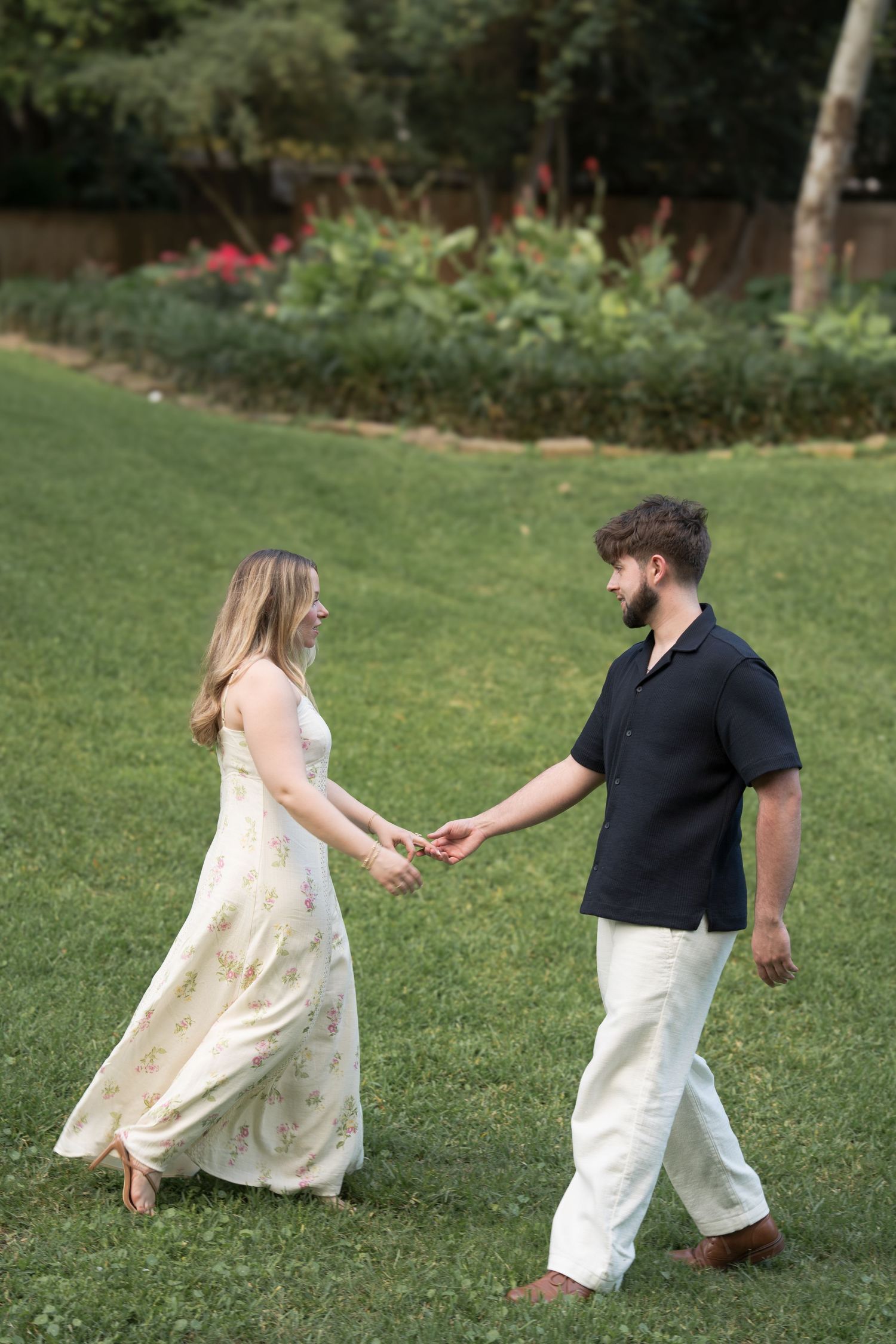 Couple holding hands and walking barefoot on green grass, woman in floral maxi dress, man in black shirt and white pants.