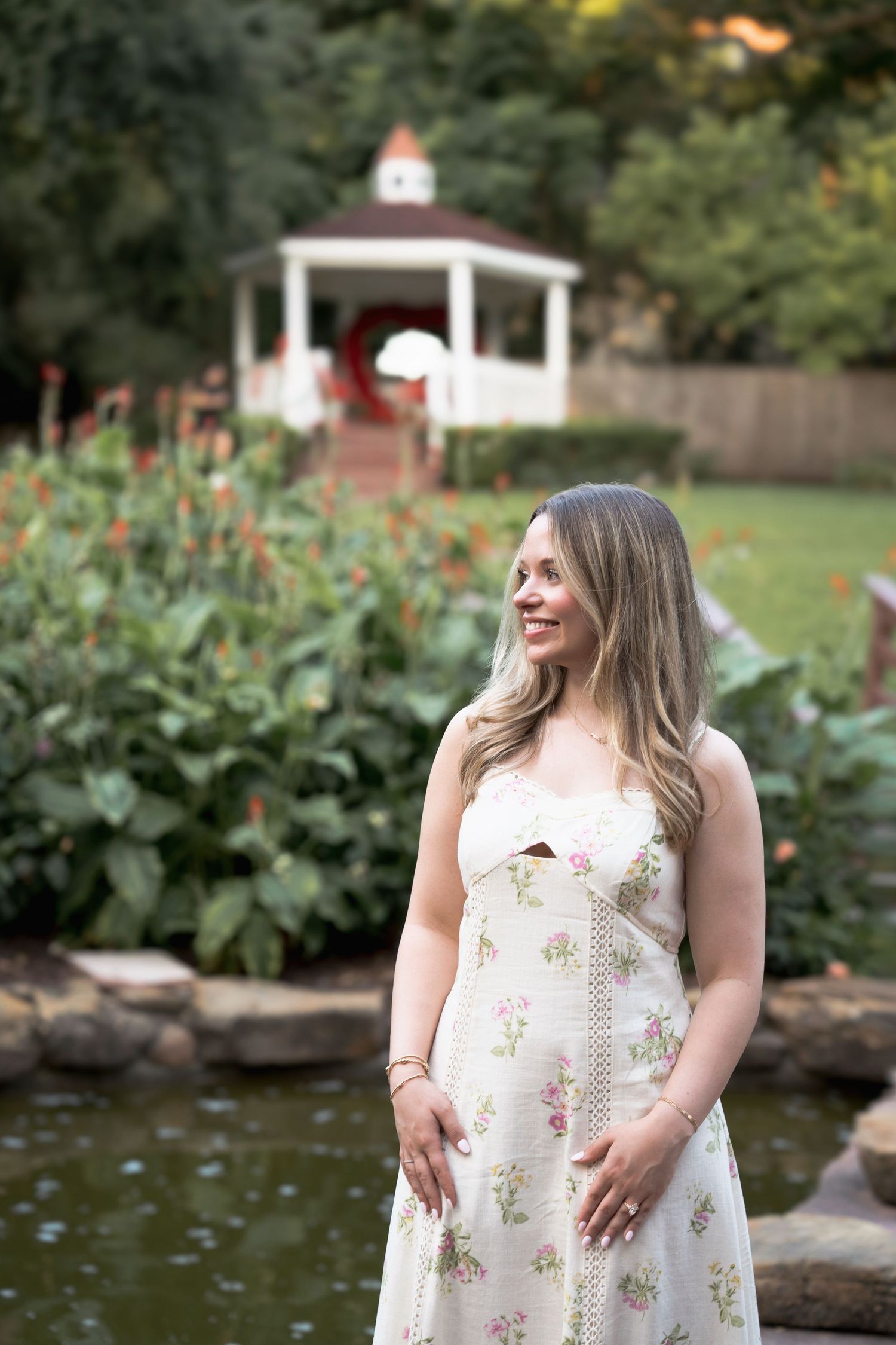 Woman in floral sundress smiling in a lush garden with a white gazebo and pond in the background.
