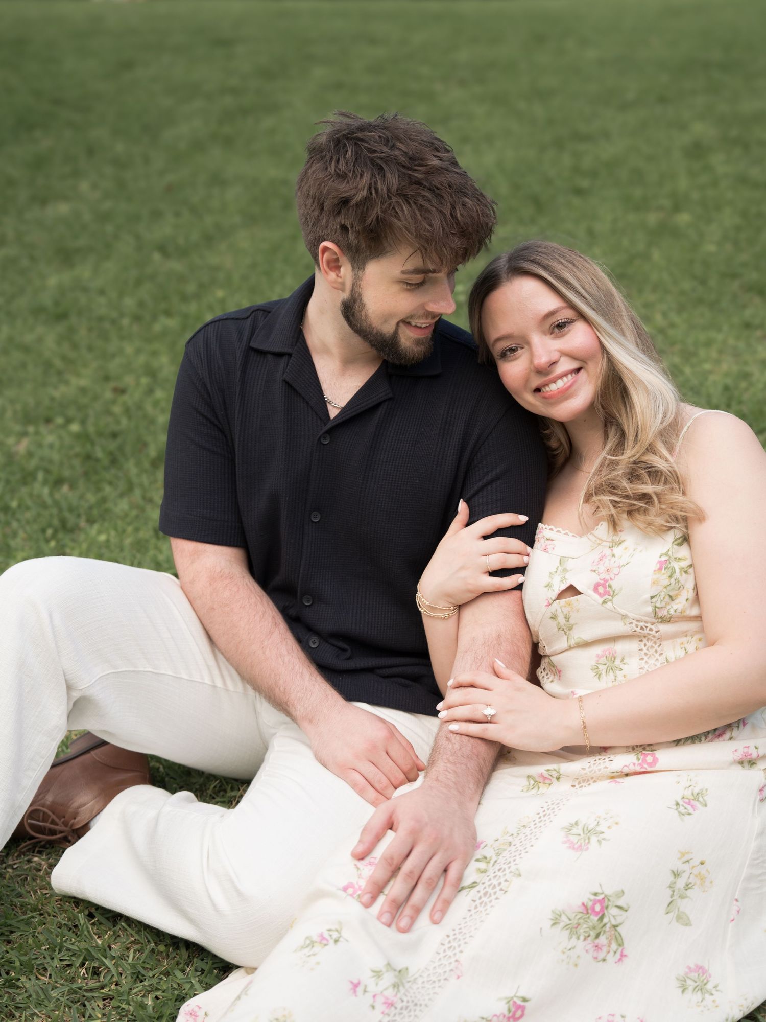 A smiling couple sits together on green grass, woman in floral dress leaning against man in black shirt and white pants.