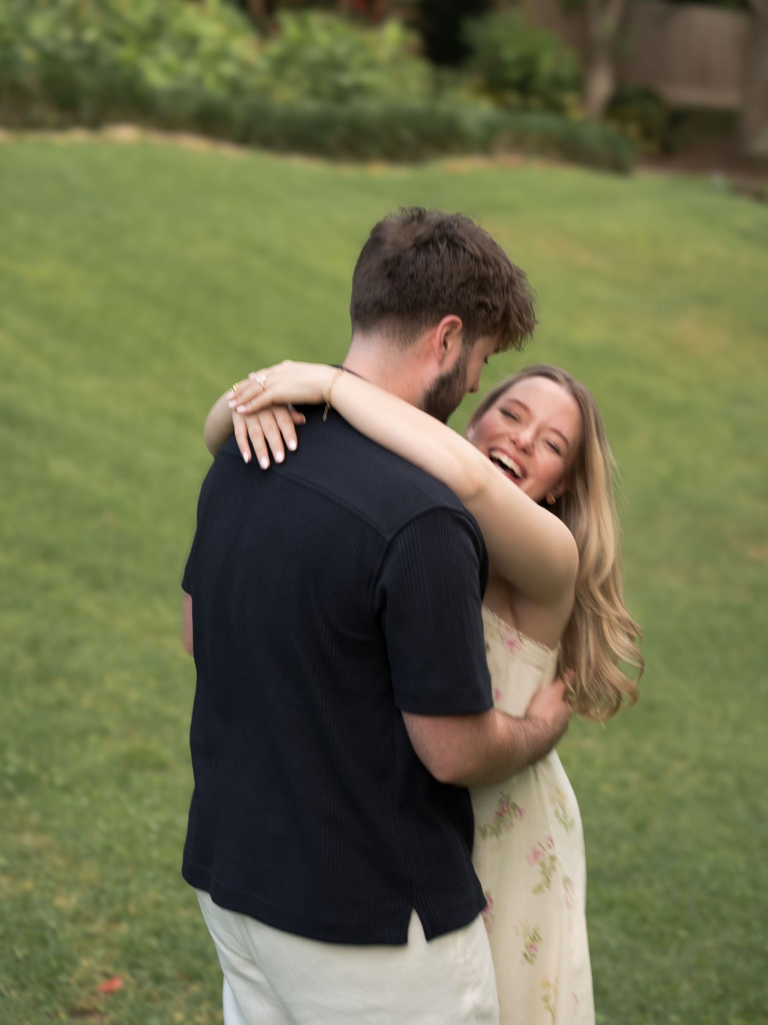 Happy couple embracing on green lawn, woman smiling joyfully while hugging man in black t-shirt outdoors.
