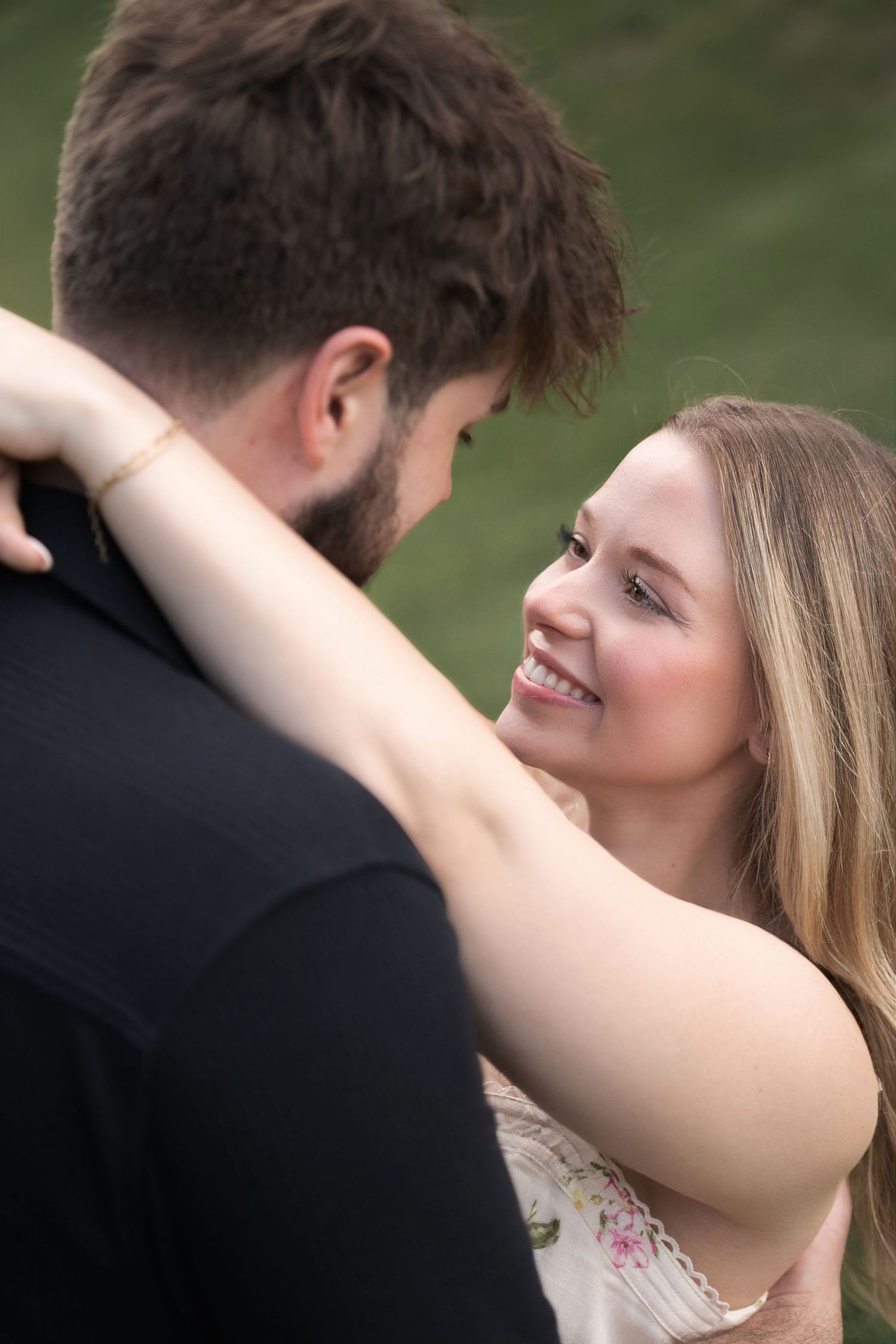 A smiling woman with long blonde hair embraces a bearded man outdoors, gazing up at him lovingly.