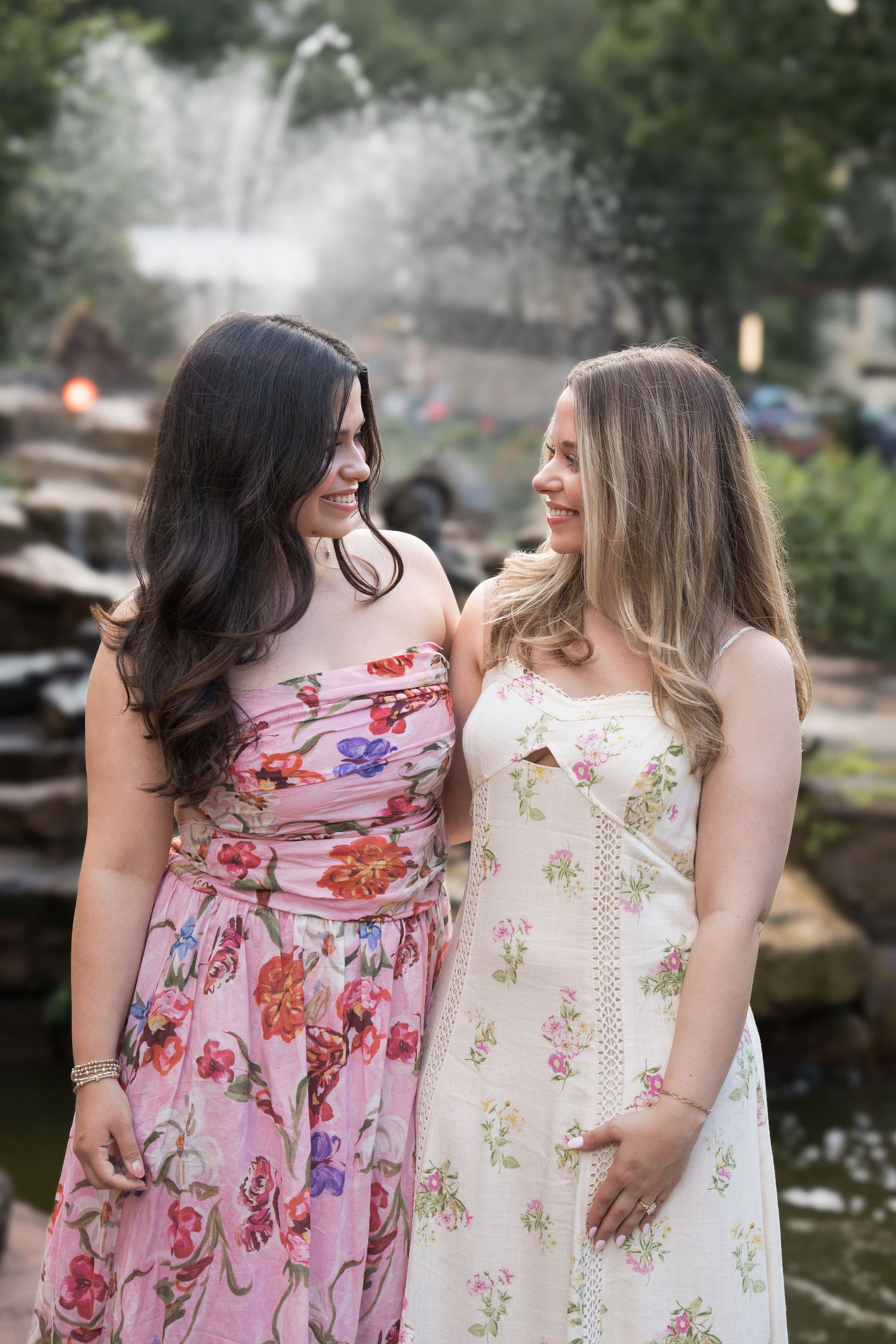 Two women in floral sundresses smile at each other near a fountain in a park.
