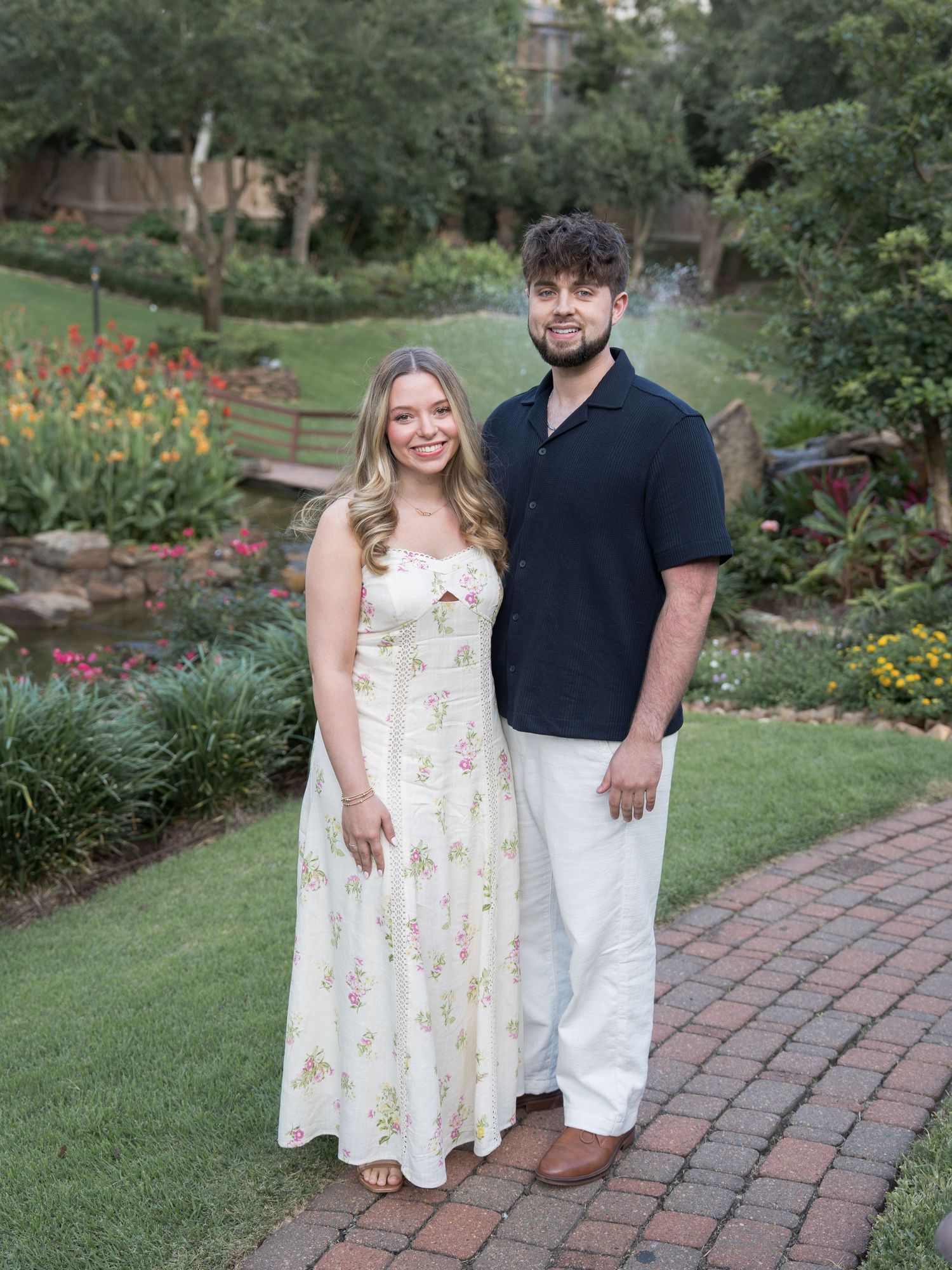 Smiling couple standing on brick path in colorful garden, woman in floral sundress and man in navy shirt.