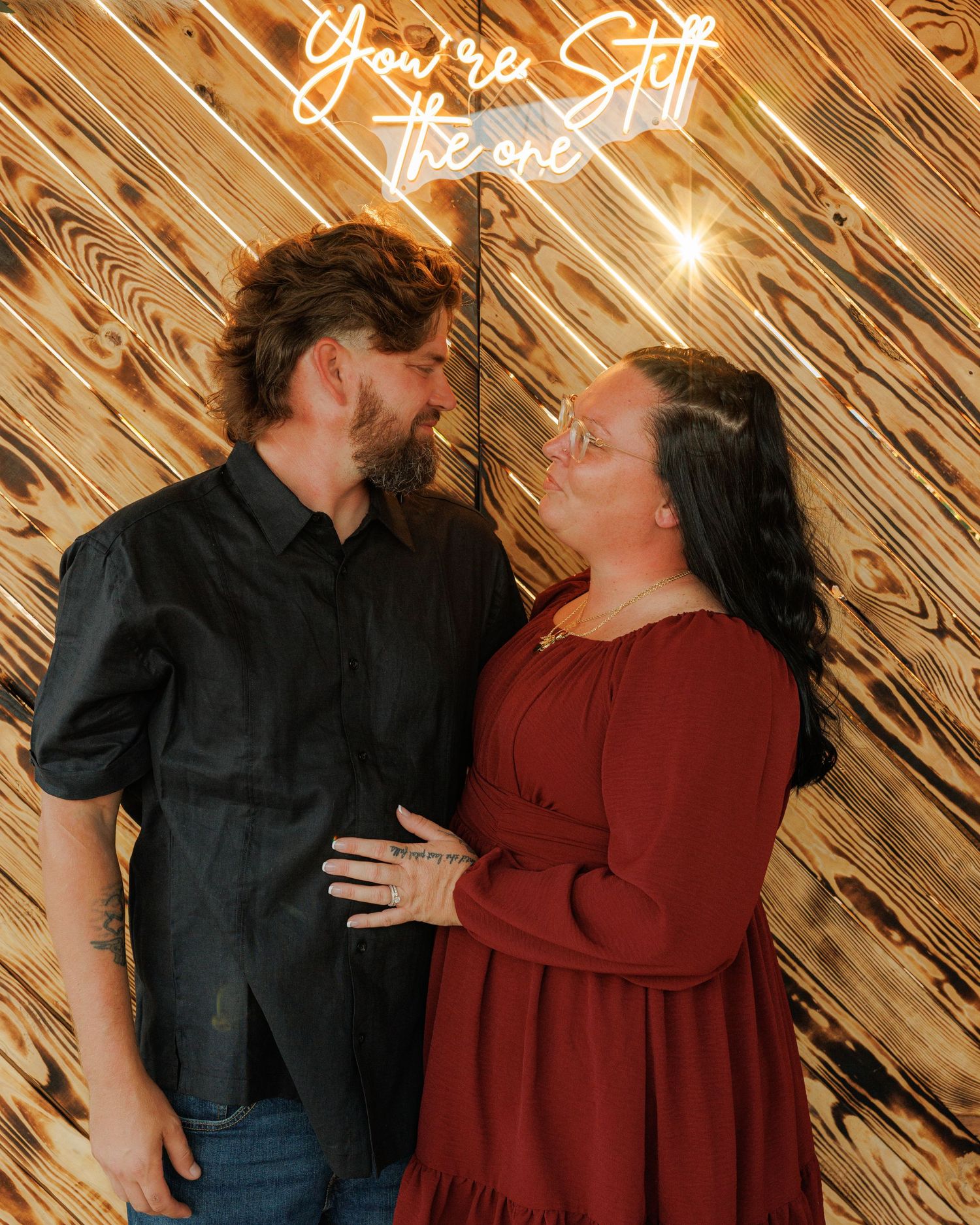 Couple sharing a romantic moment in front of a wooden wall with neon sign reading You're Still The One.