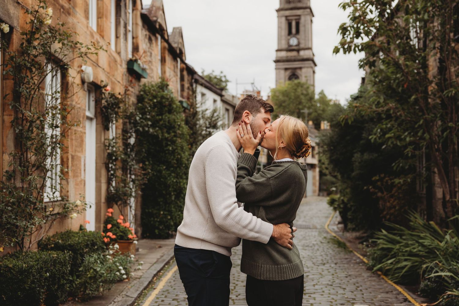 Couple sharing a romantic kiss on a cobblestone street in Edinburgh, Scotland, with historic stone buildings and a church tower.