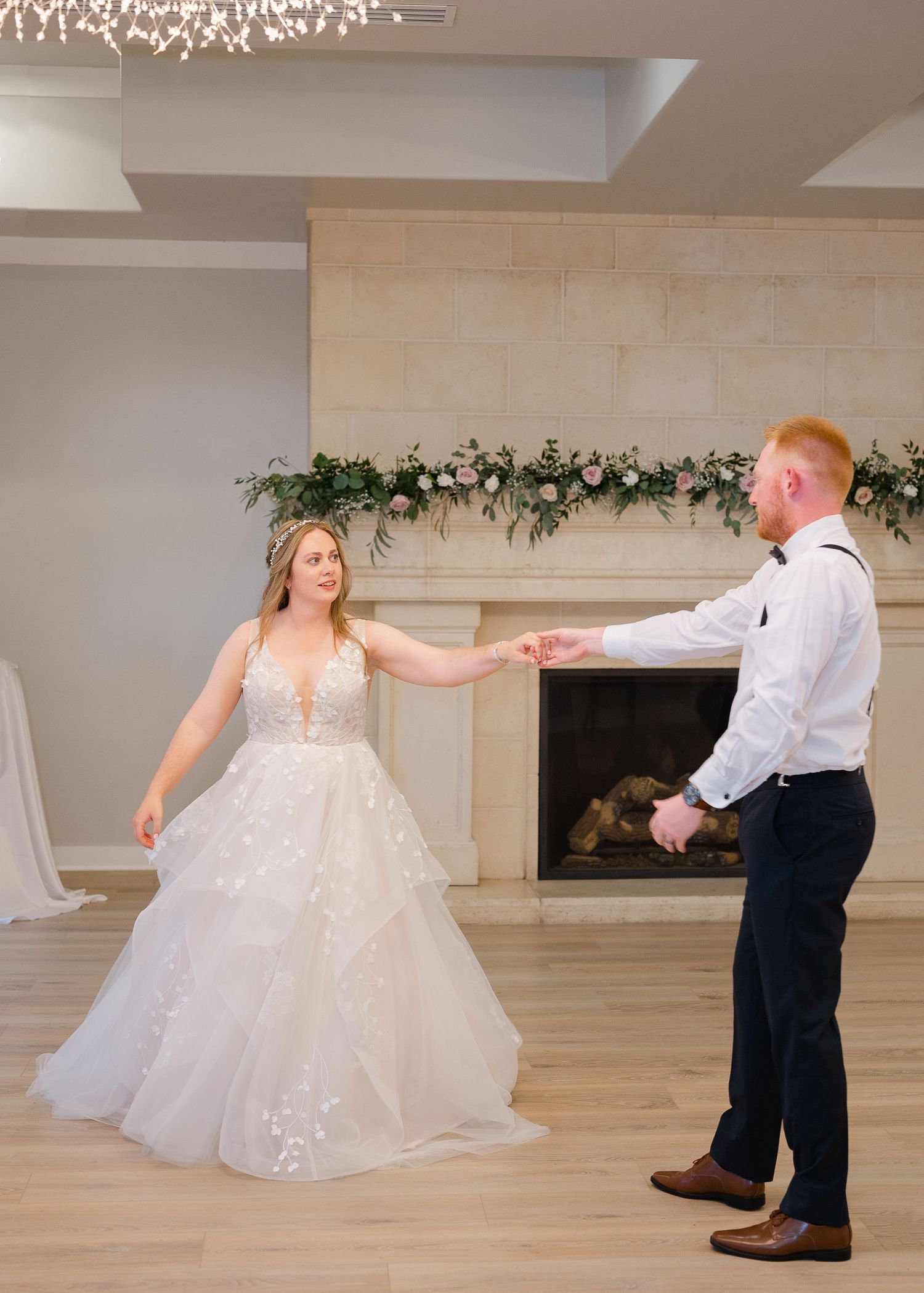 Bride and groom share their first dance near a floral-adorned fireplace in an elegant wedding reception venue.