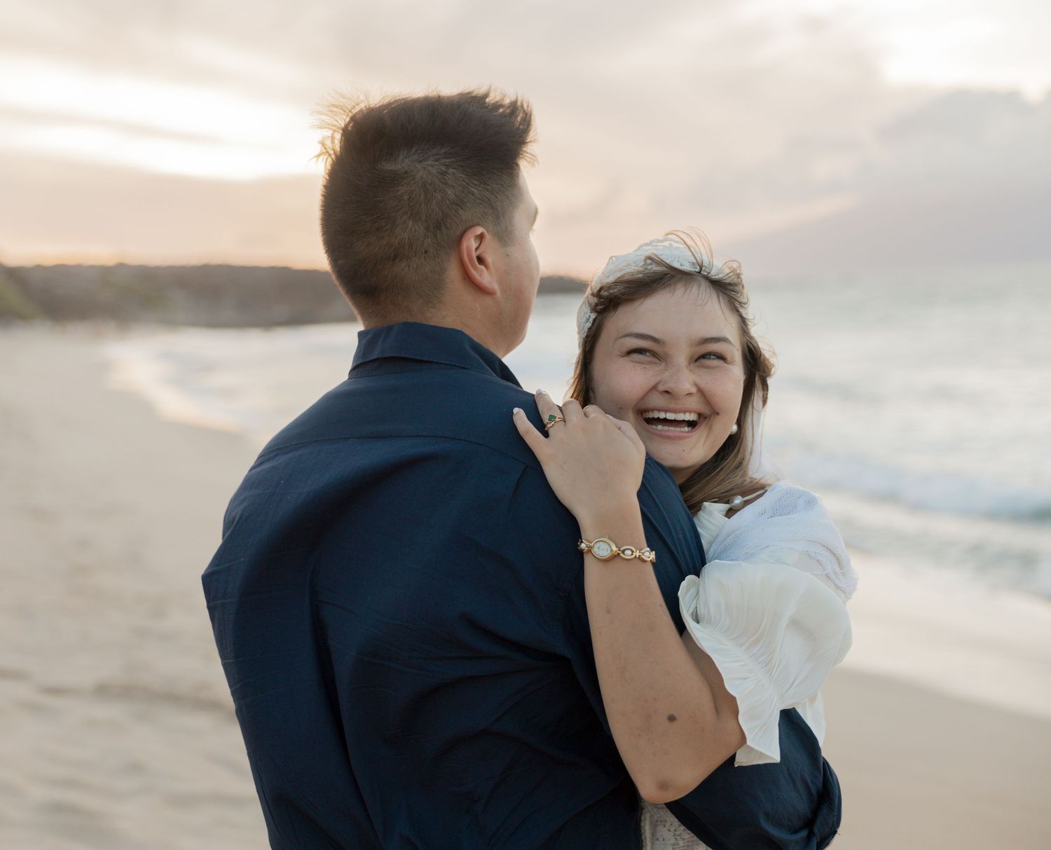 Happy couple embracing on a beach at sunset, woman laughing joyfully in white dress, man in navy suit.