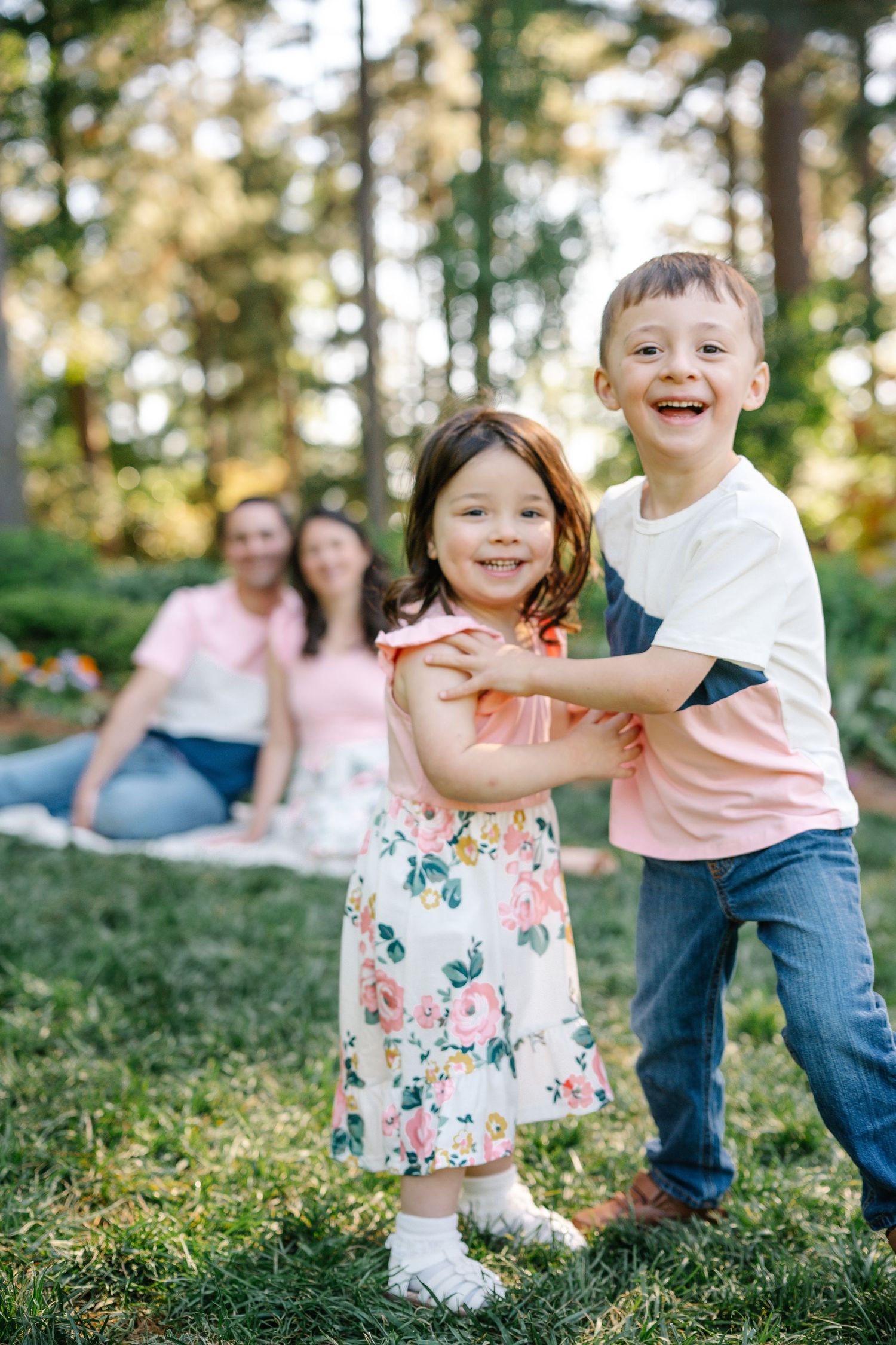 Two happy children playing in a sunny park while their parents relax on the grass in the background.