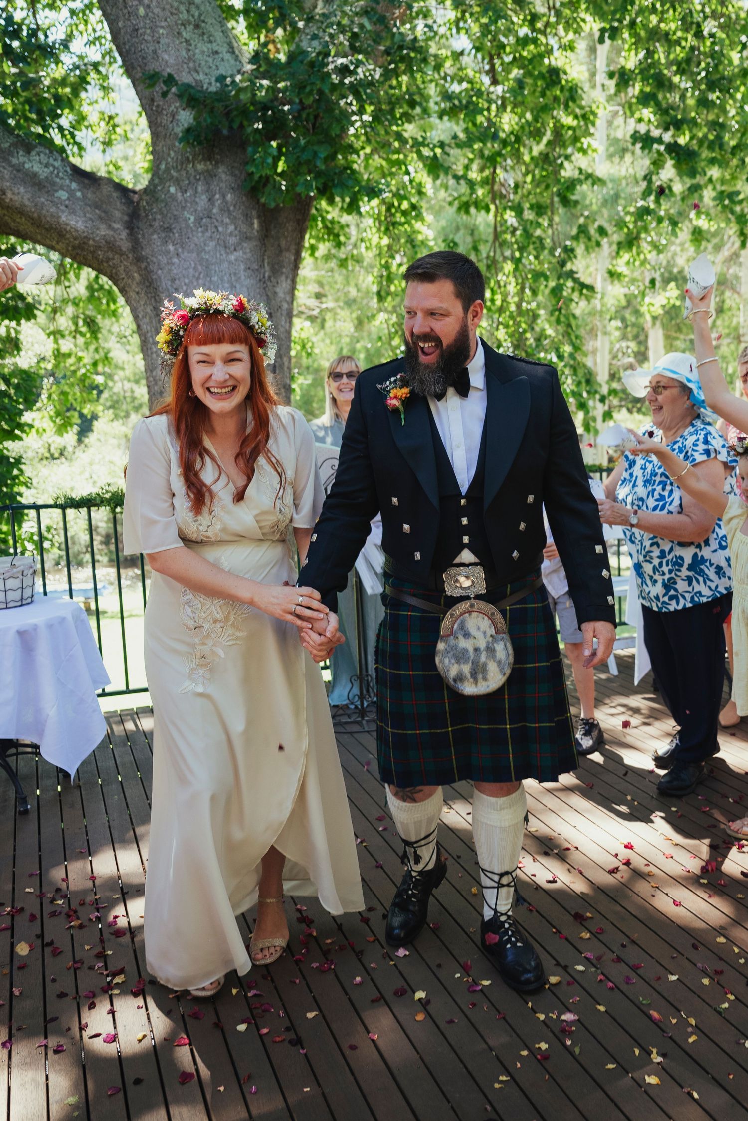 Bride in white dress with flower crown and groom in Scottish kilt laughing together at outdoor wedding ceremony.