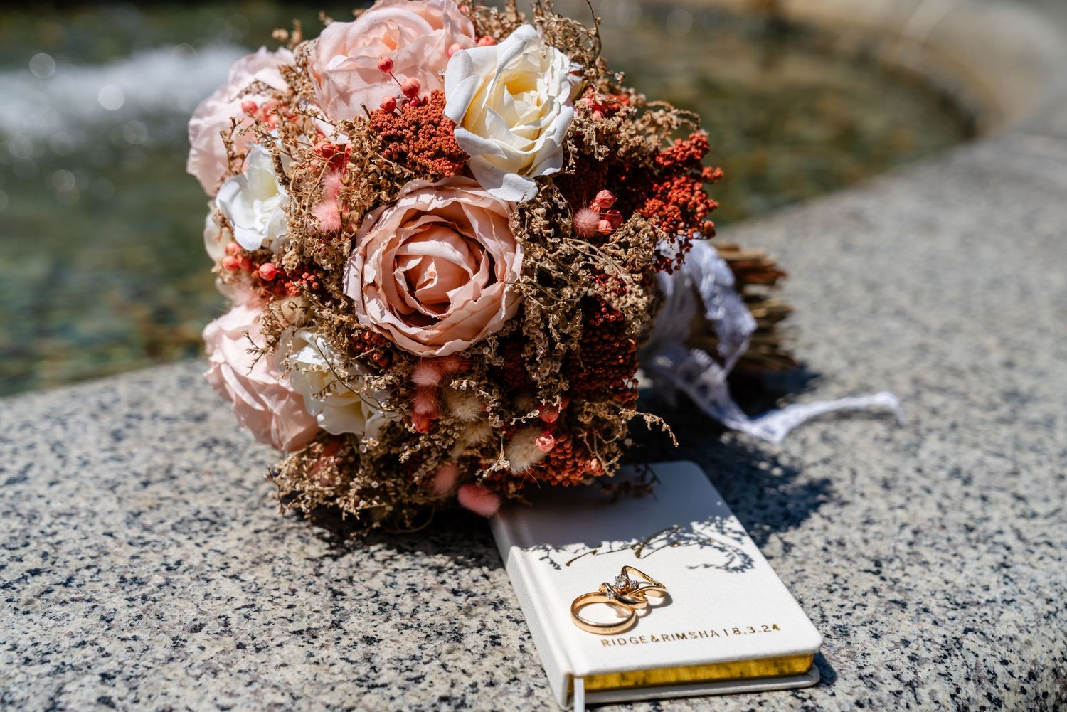 Wedding bouquet of pink roses and berries resting on a book with gold rings beside a fountain.