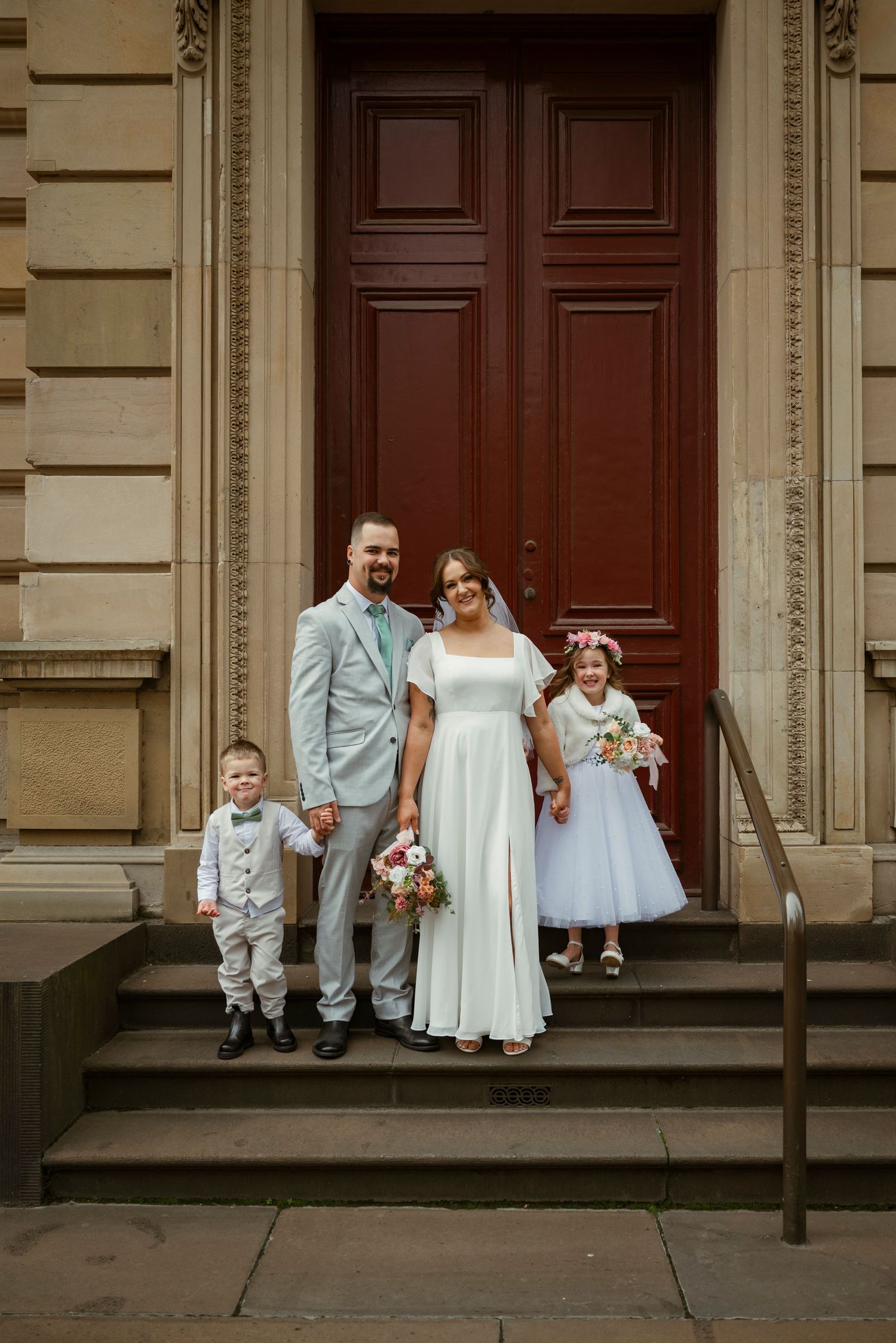 Wedding family portrait on church steps, bride in white gown, groom in grey suit, with ring bearer and flower girl.