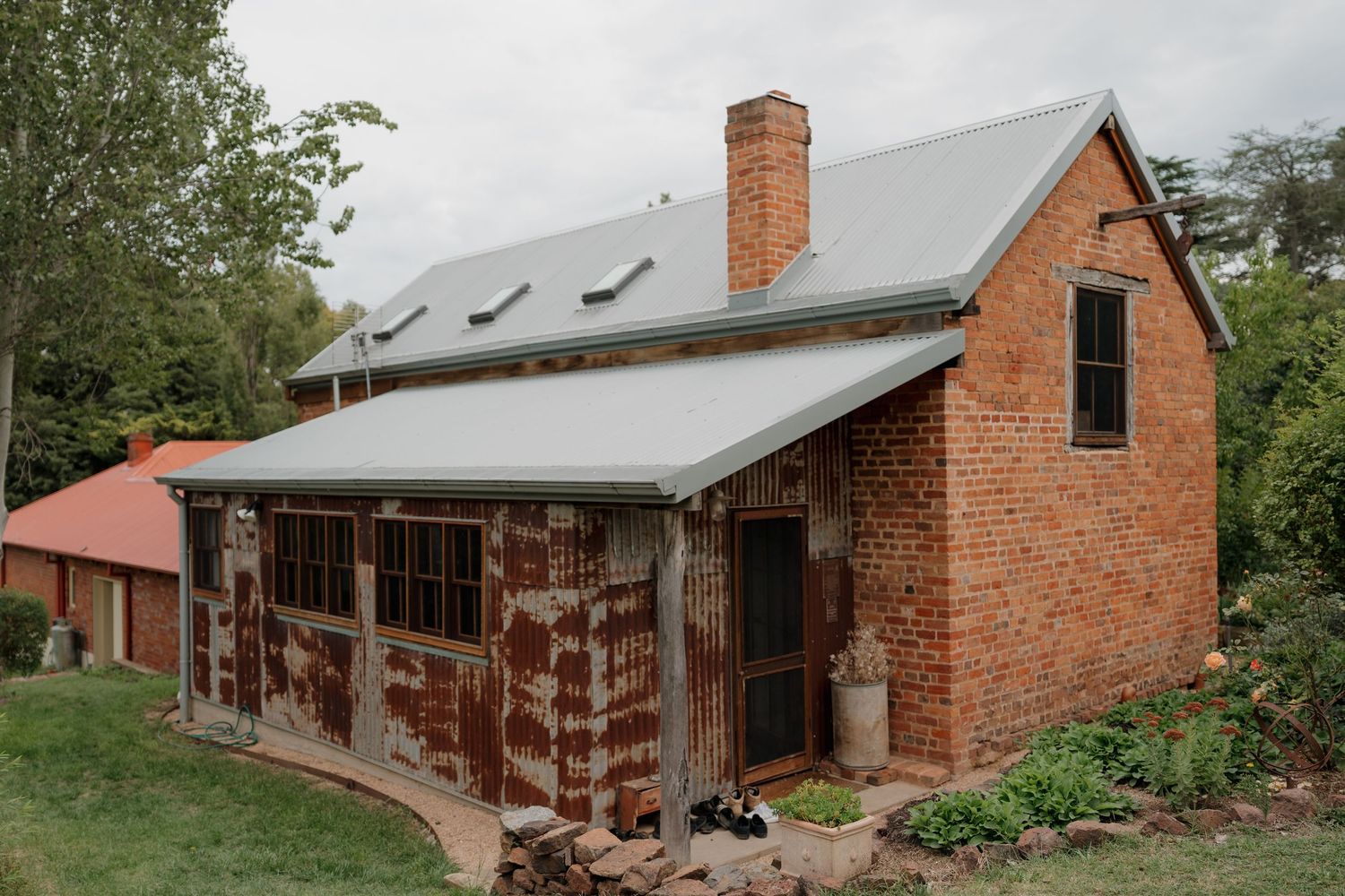 Rustic red brick cottage with grey metal roof, chimney, and lush green surroundings in a rural Australian setting.