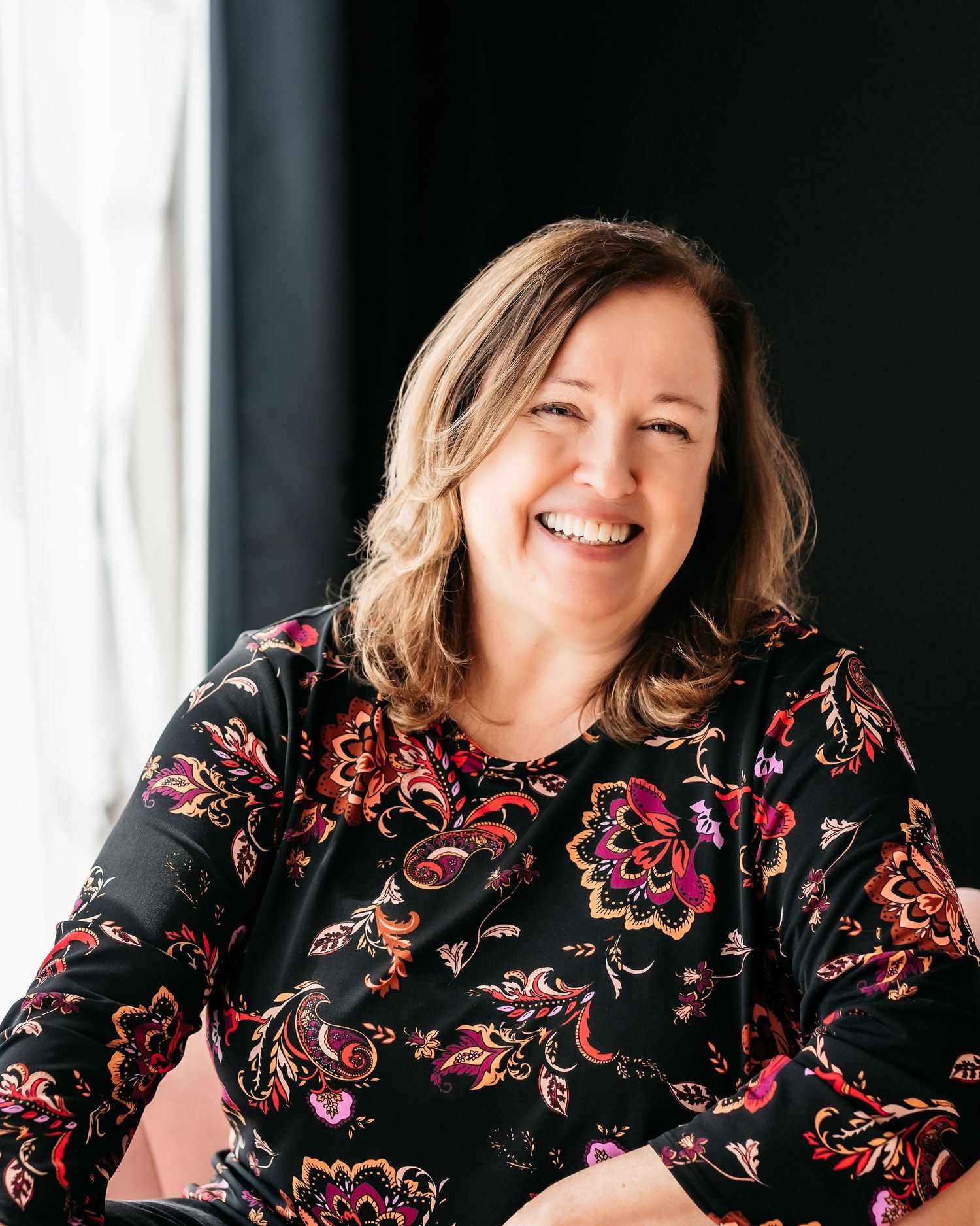 Smiling middle-aged woman with shoulder-length hair wearing a floral black dress, seated in a bright modern setting.