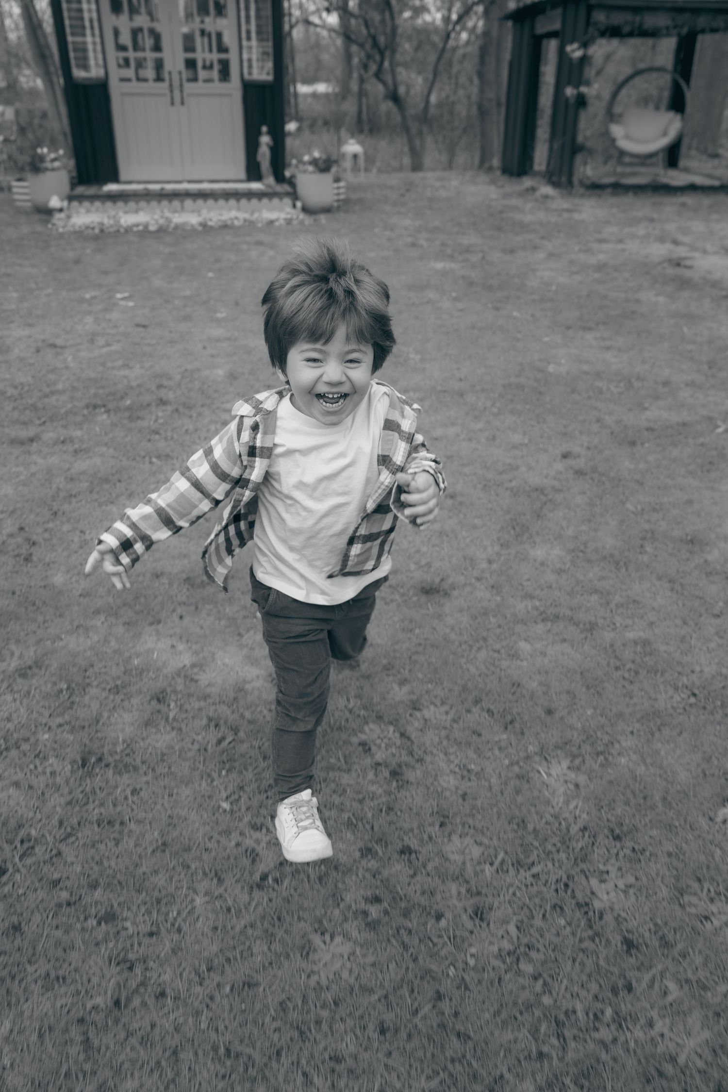 Black and white photo of a joyful young boy running and laughing in a backyard garden near a small shed.