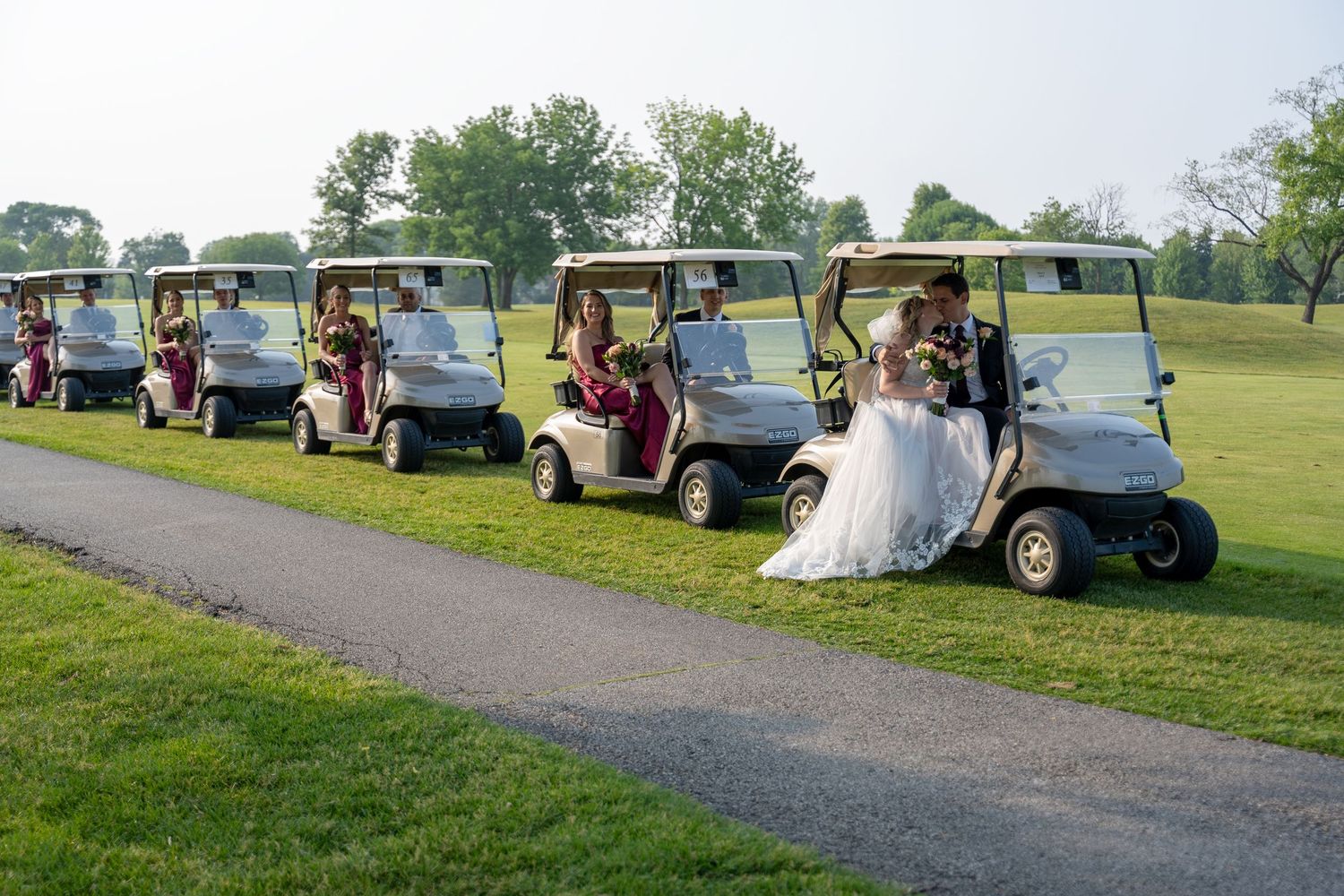 Wedding party rides golf carts across a lush green golf course, bride and groom leading in white.