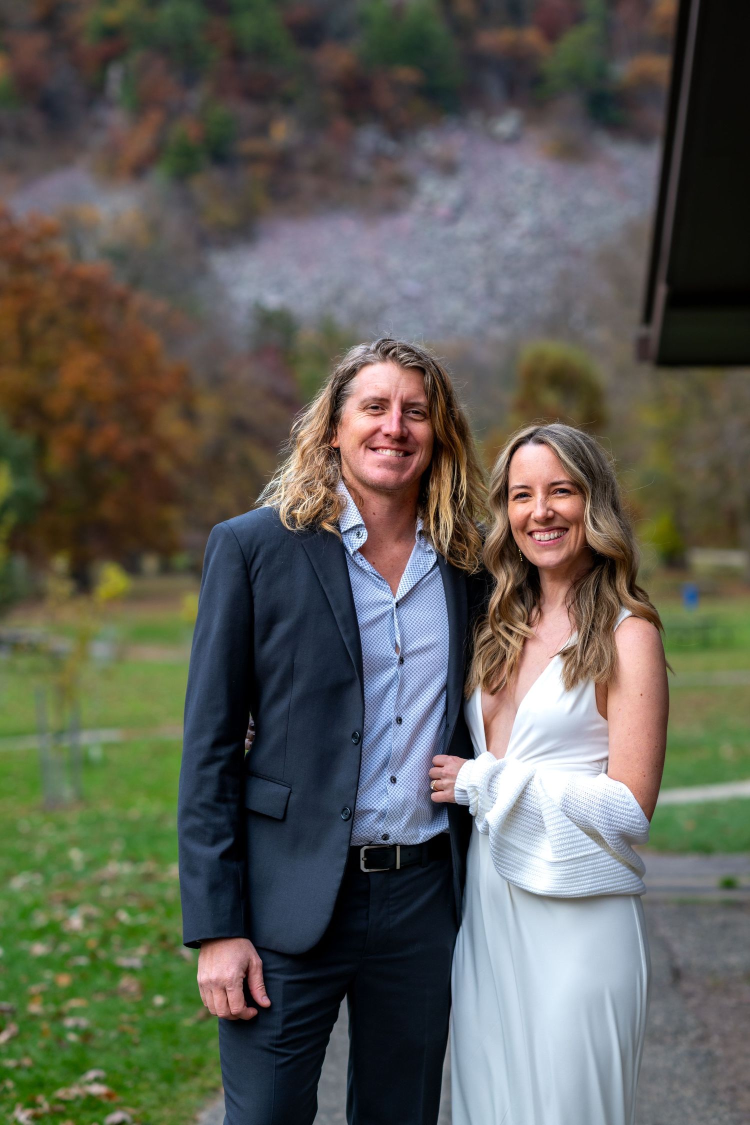 Smiling couple in wedding attire posing outdoors with autumn foliage in background.
