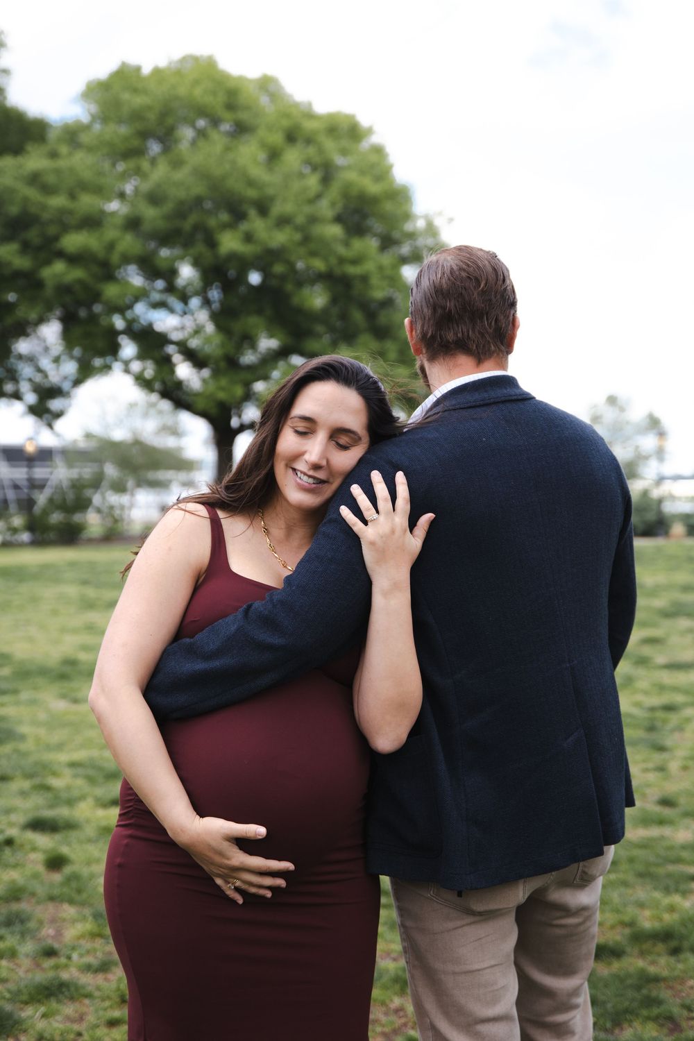 Pregnant woman in burgundy dress smiling with eyes closed, leaning against partner in navy blazer outdoors.