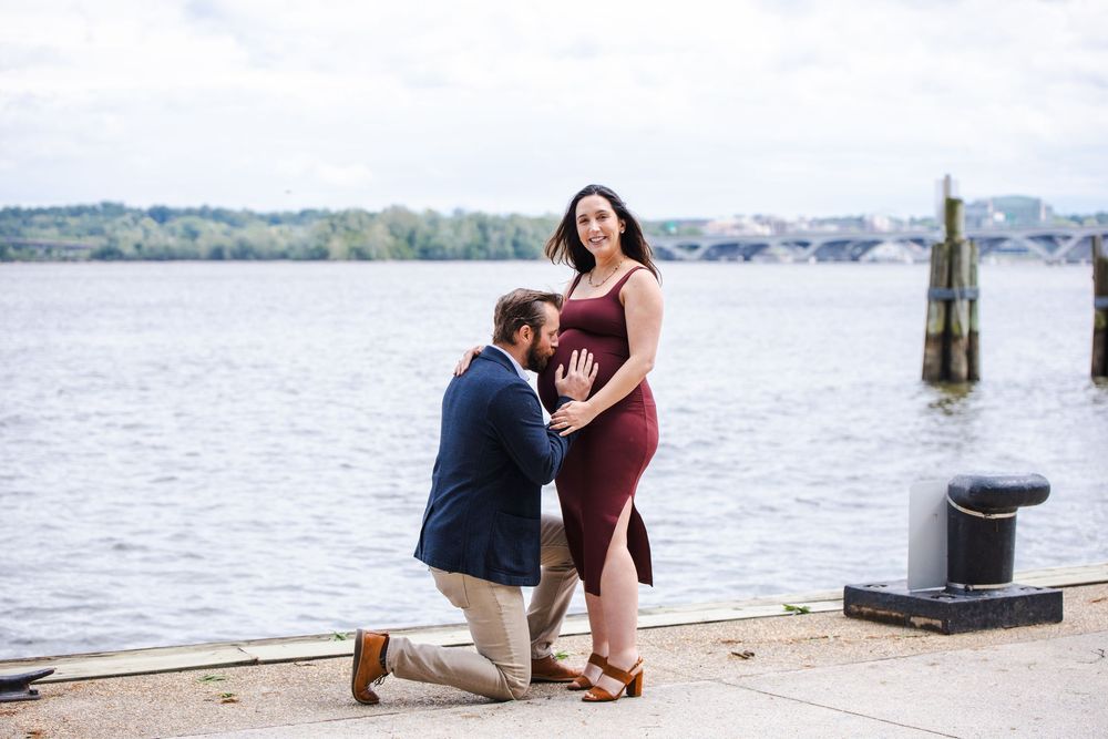 Man on one knee proposing to a woman by the waterfront, she shows her ring while he kisses her hand.
