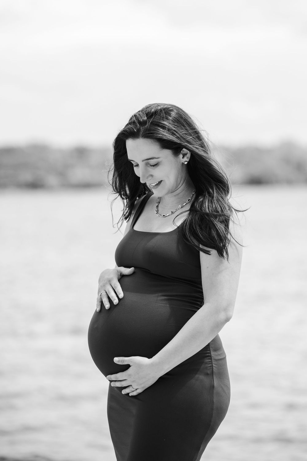 Black and white photo of a pregnant woman cradling her baby bump near the water, smiling softly downward.