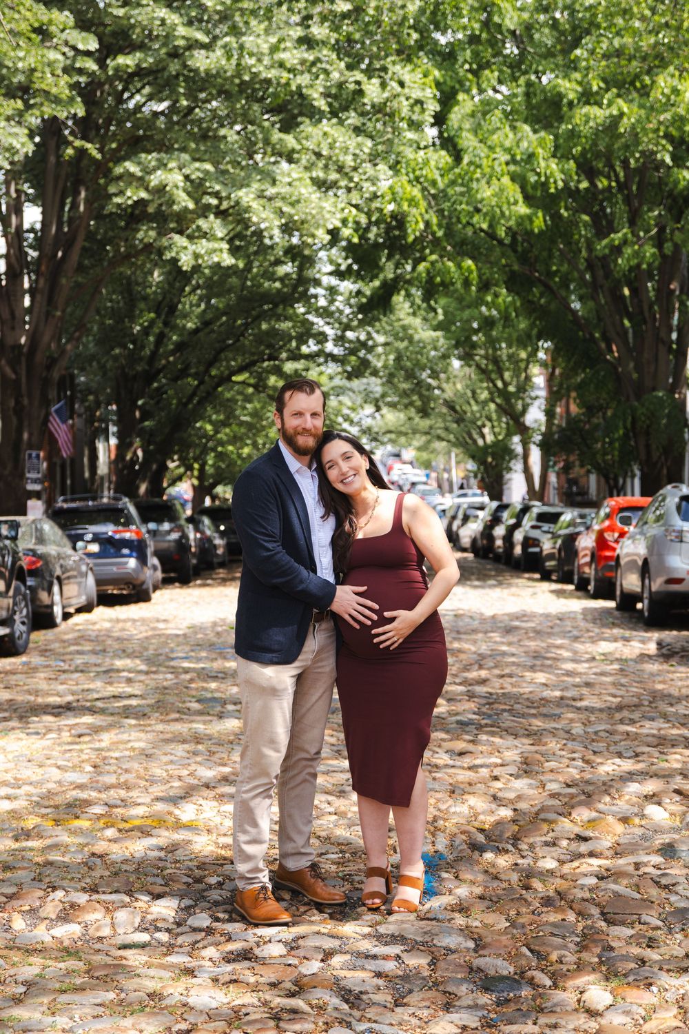 Couple poses on cobblestone street, woman in burgundy maternity dress, man in navy blazer, tree-lined urban setting.