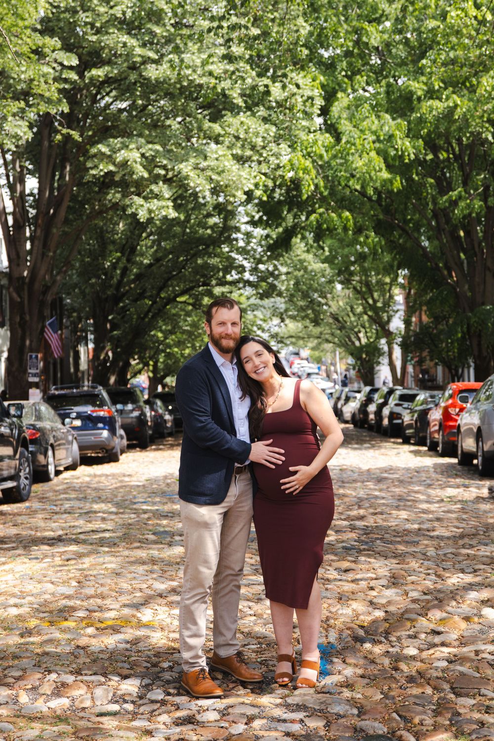 Pregnant couple posing on a cobblestone street lined with trees and parked cars, man in blazer embracing woman in burgundy dress.