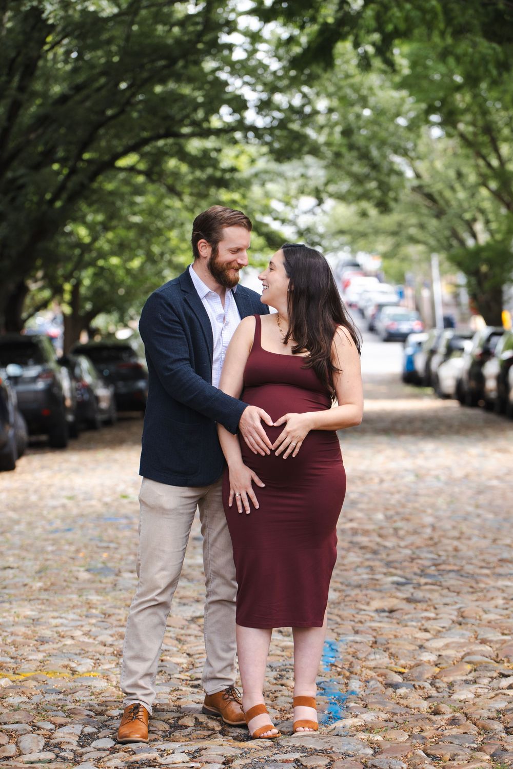Couple making heart shape on pregnant belly on a tree-lined cobblestone street.