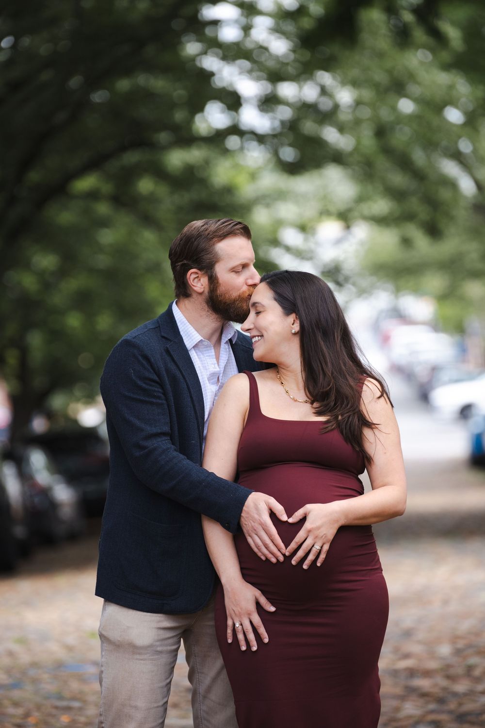 Couple sharing a kiss on a tree-lined cobblestone street, man forming heart shape on pregnant partner's belly.
