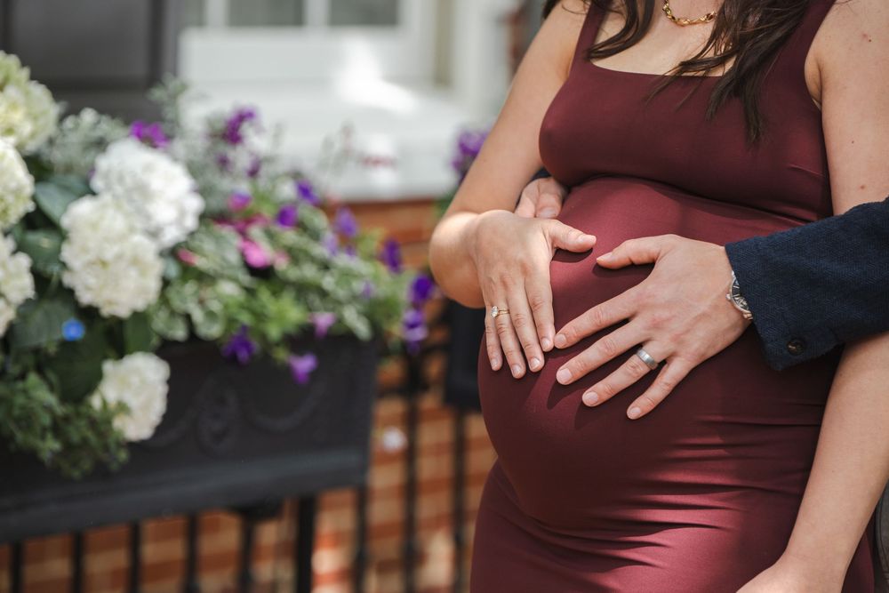 Couple tenderly cradling pregnant belly in burgundy dress outdoors near white and purple flowers.