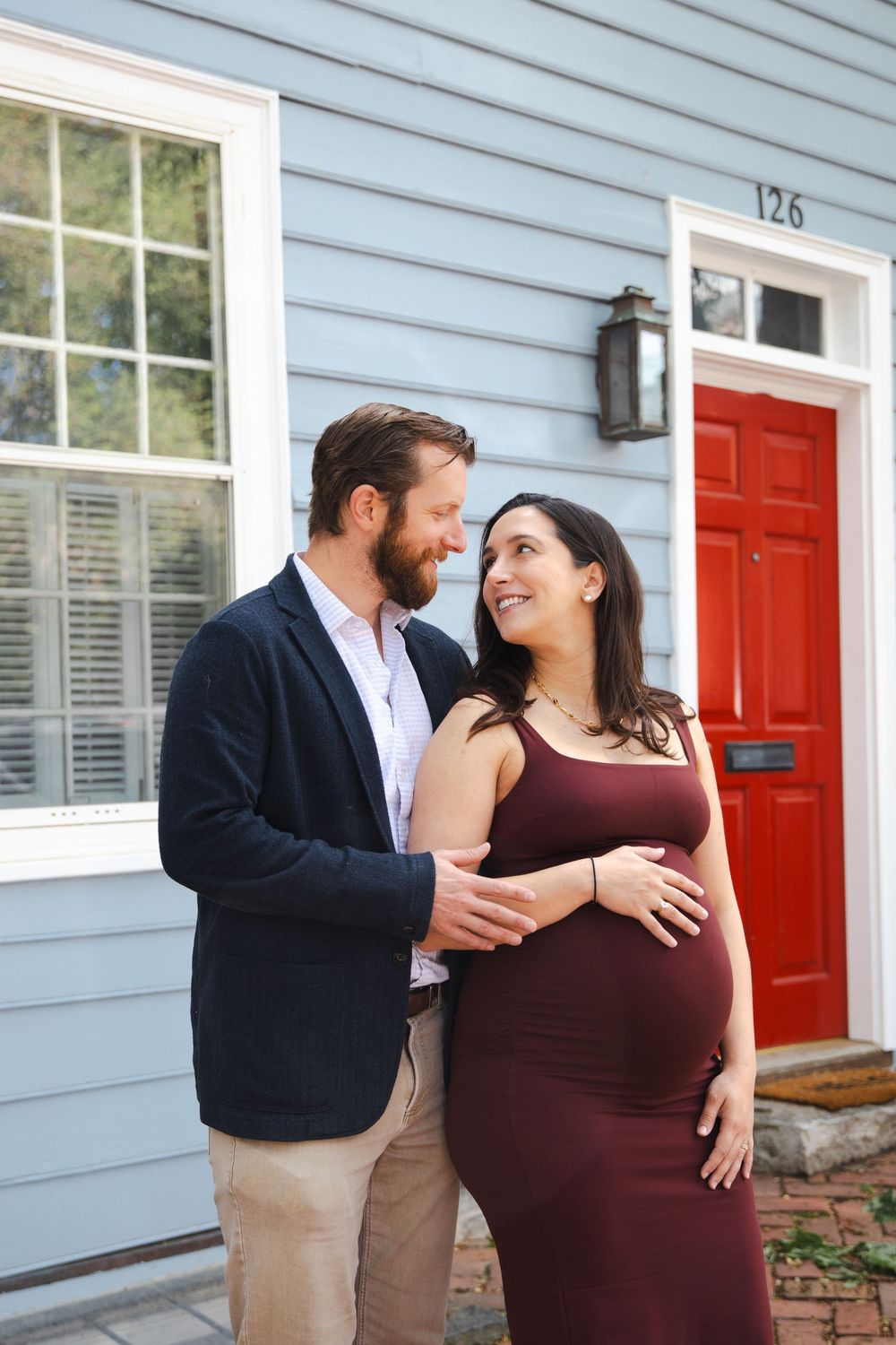 Smiling couple sharing a tender moment outside a blue house with red door, pregnant woman in burgundy dress.