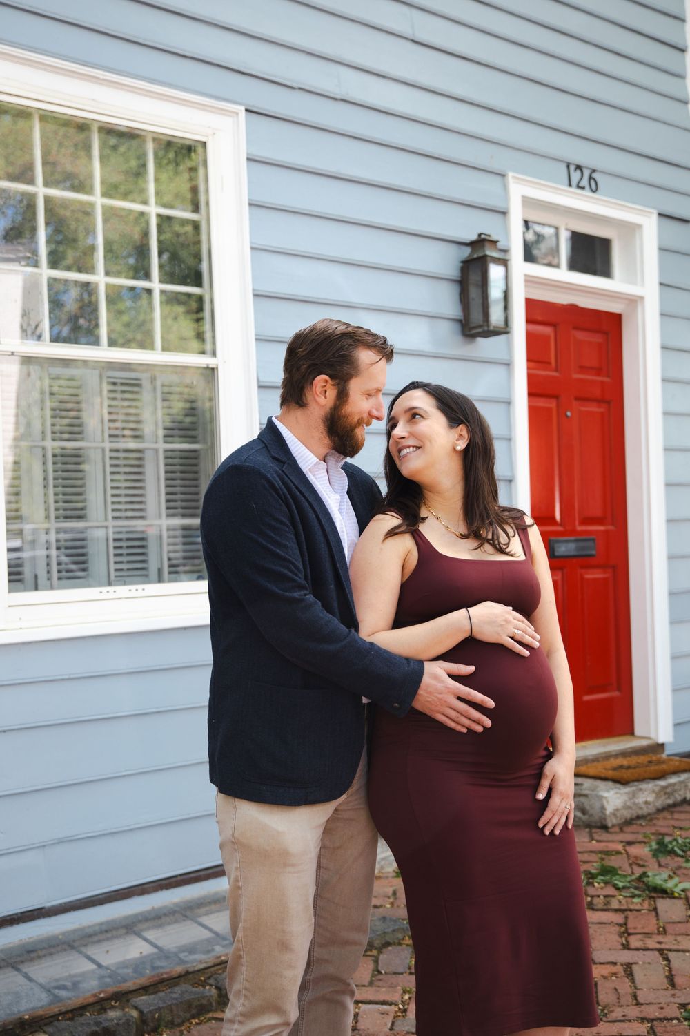 Smiling couple shares tender moment outside blue house with red door, woman in burgundy maternity dress.