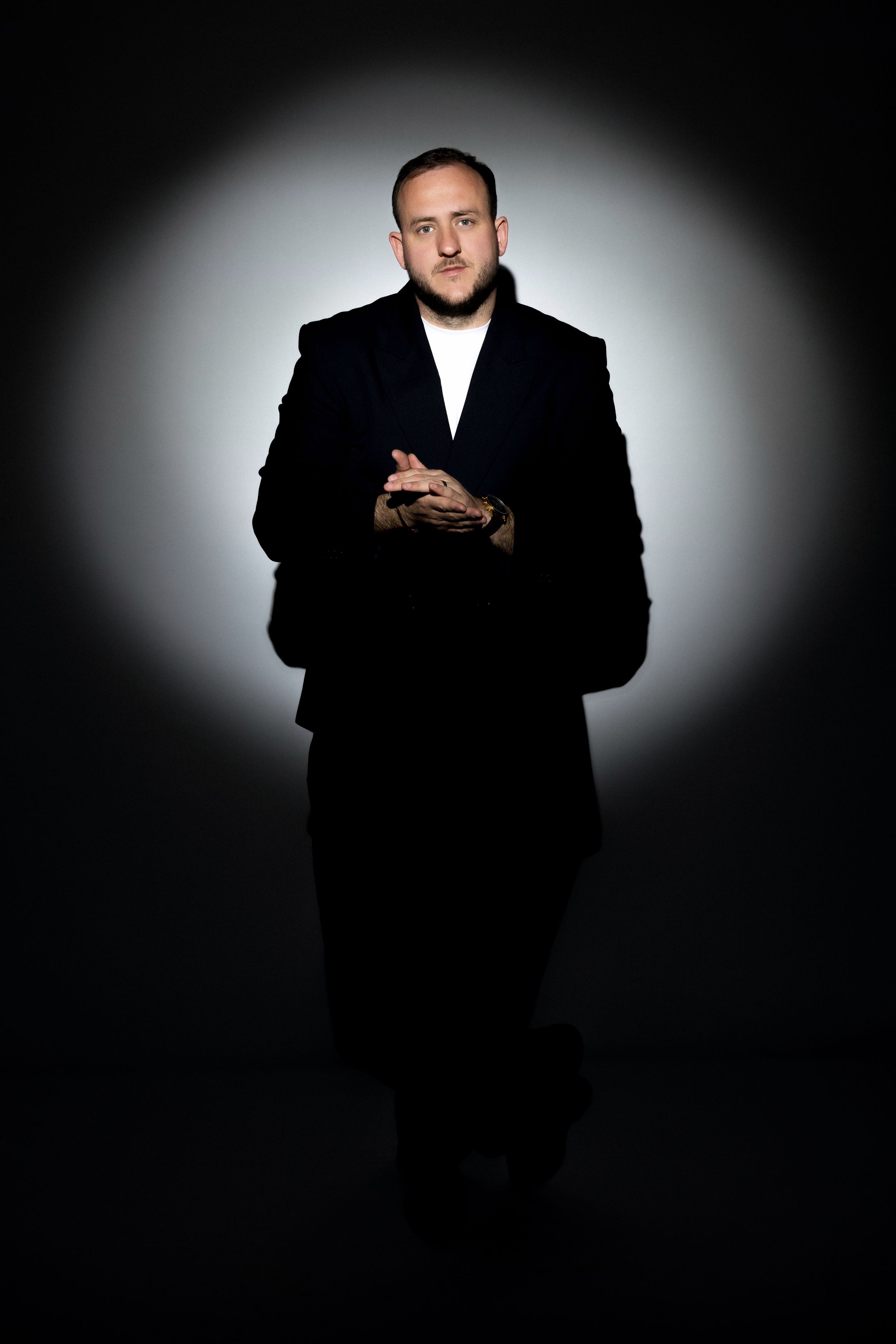 Man in black coat with tattoos stands confidently against dramatic spotlight background.