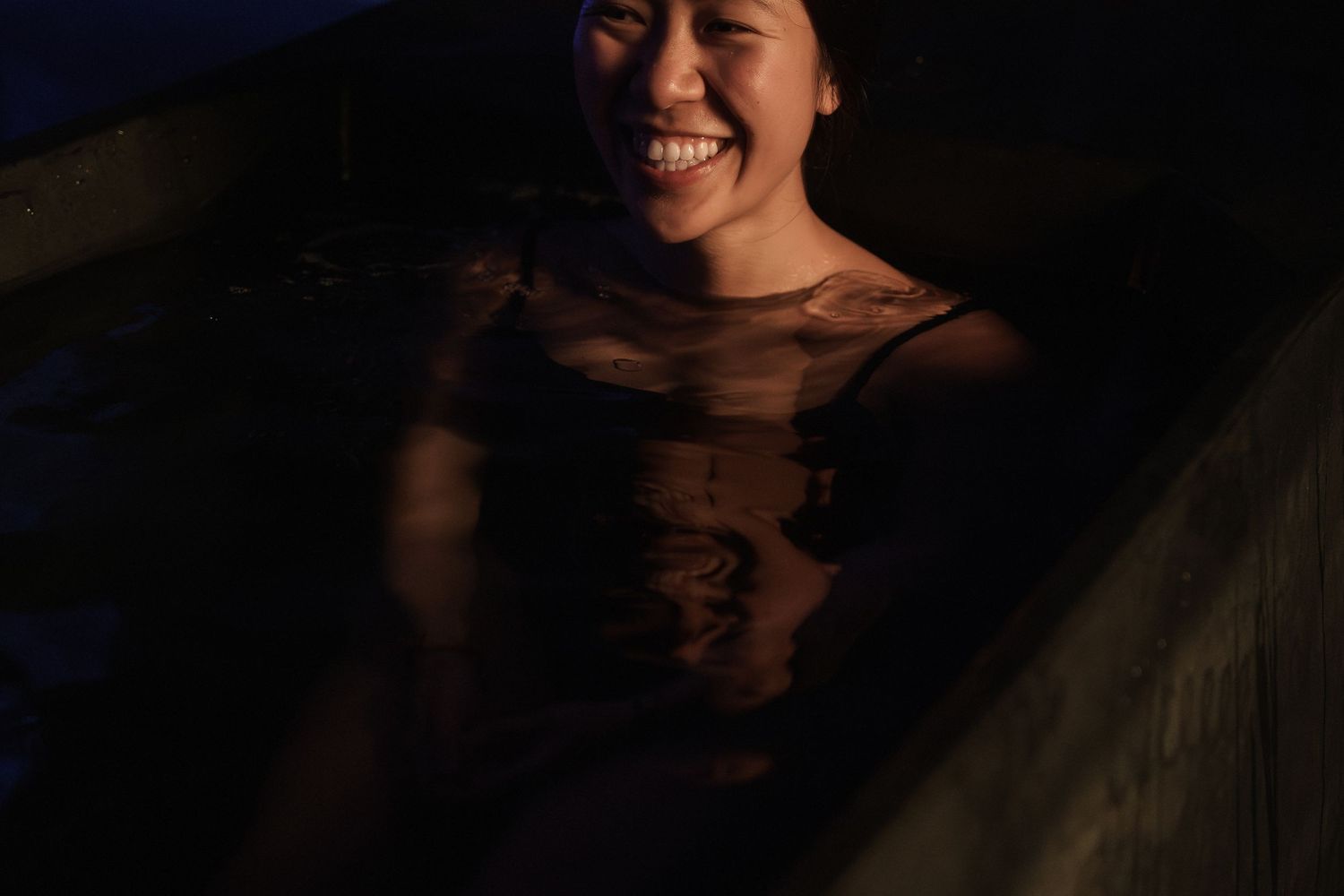 Smiling woman relaxing in a dark outdoor hot spring bath at night, enjoying a peaceful soak.