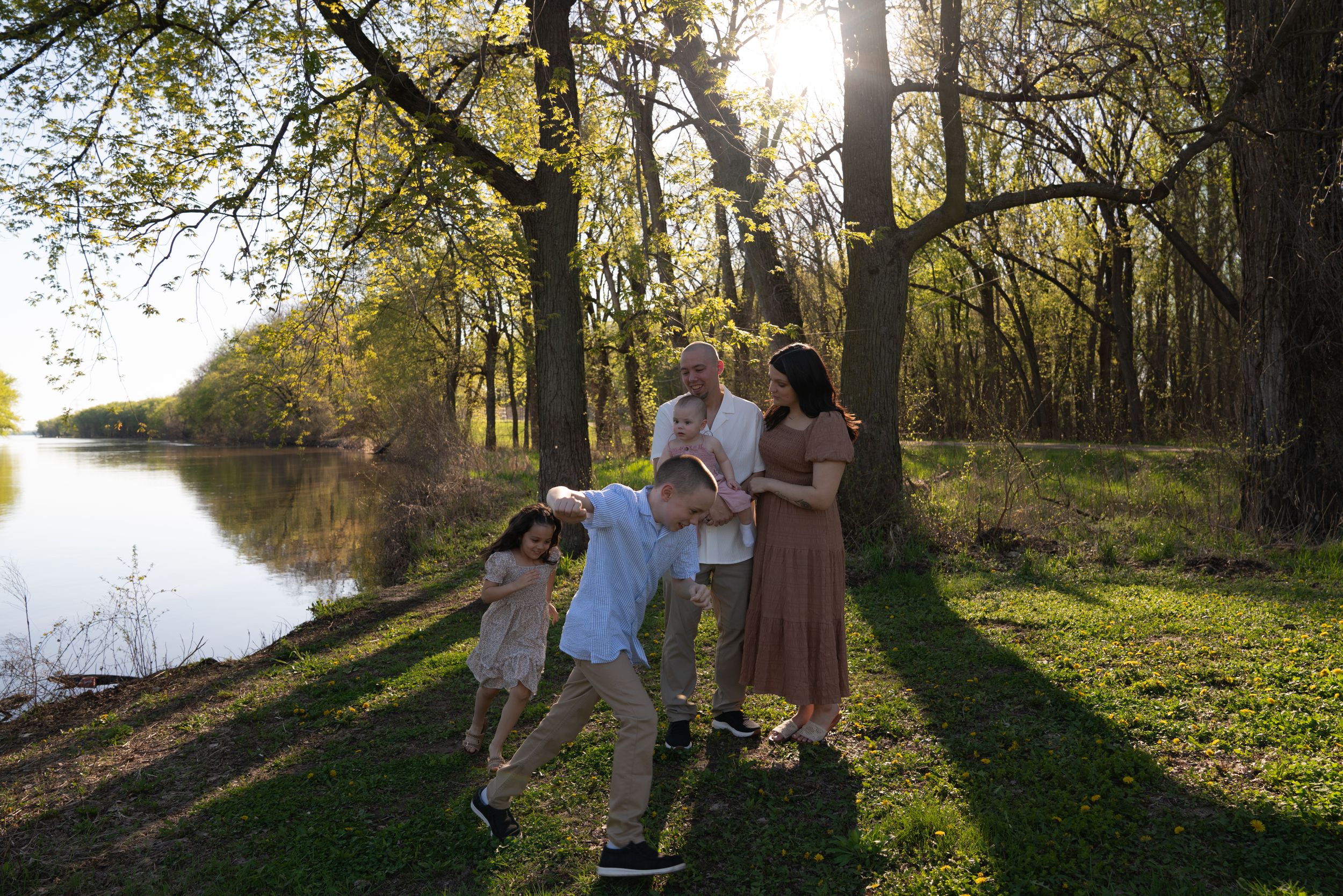 Family of five enjoying a sunny day by a river, with children playing and parents smiling among lush green trees.