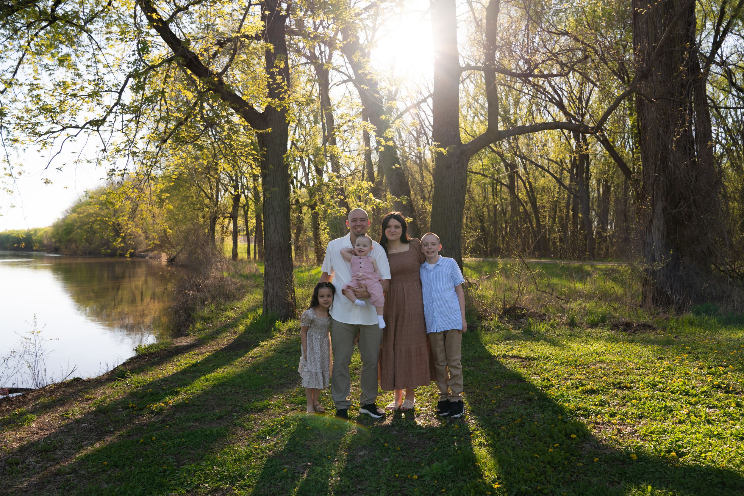 Family of five posing together near a sunlit pond surrounded by tall trees during golden hour outdoors.