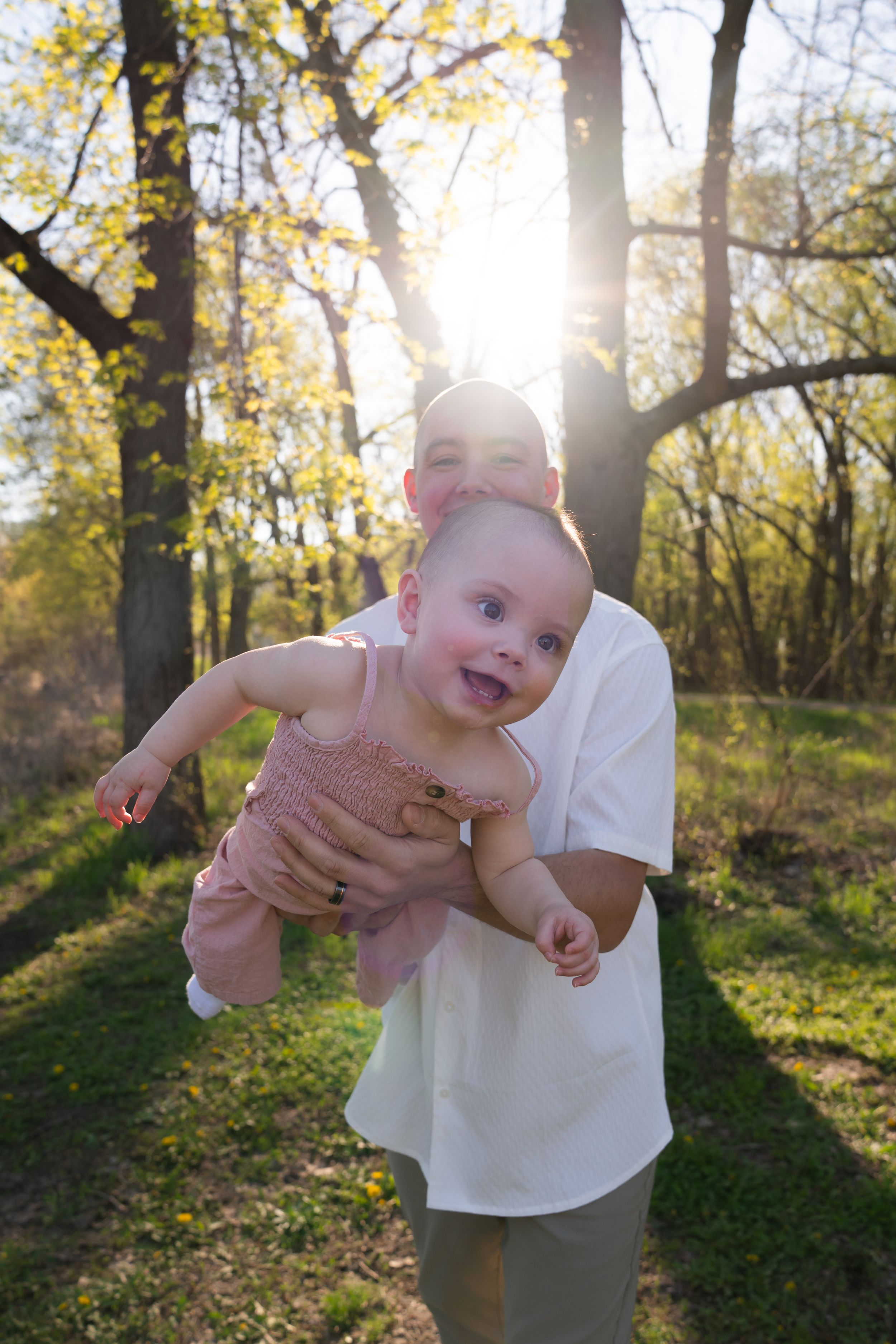 Parent holding a happy baby outdoors in sunlit forest during golden hour, baby wearing pink outfit.