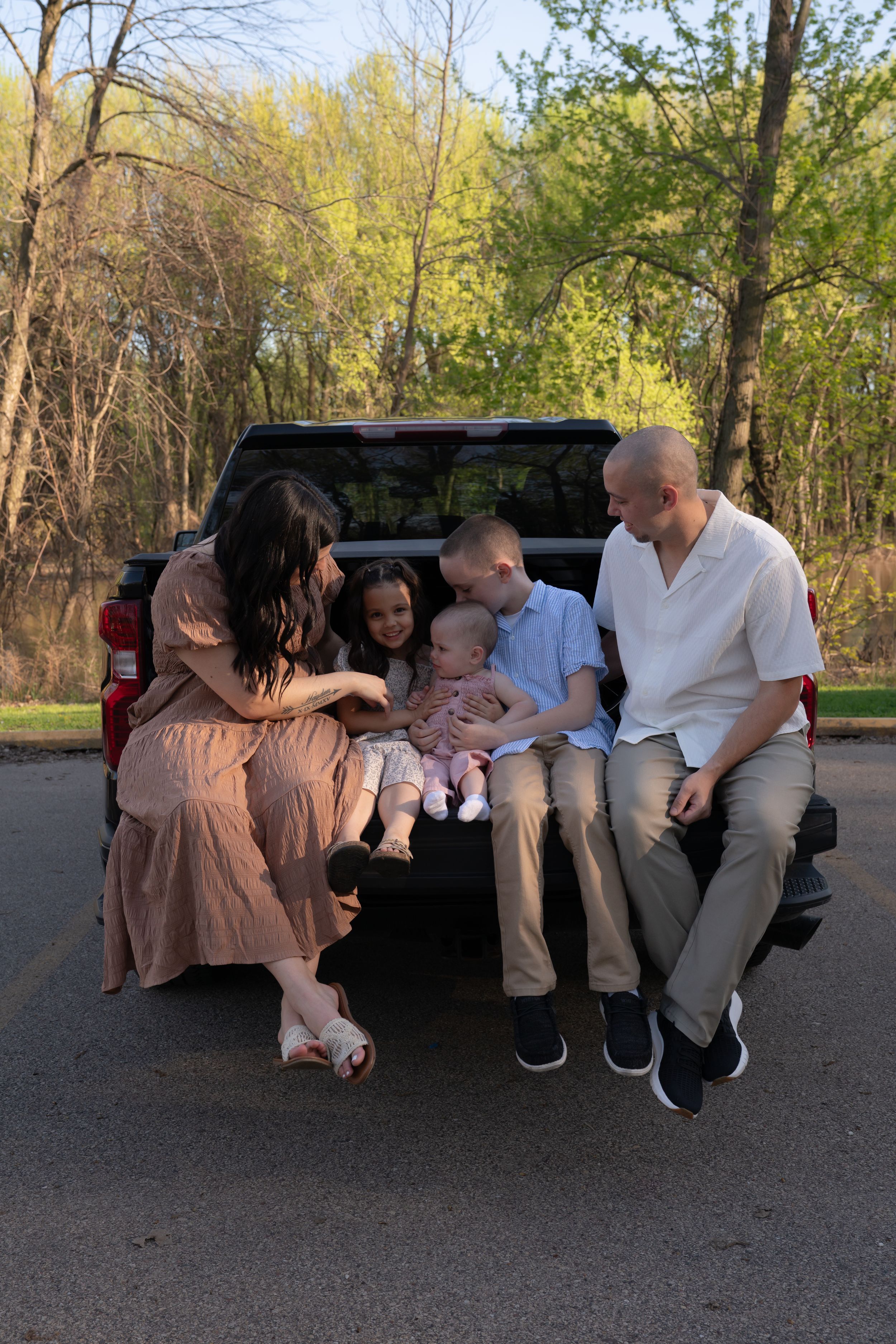 Family of five sitting on truck tailgate outdoors, parents with two children and baby in spring setting.