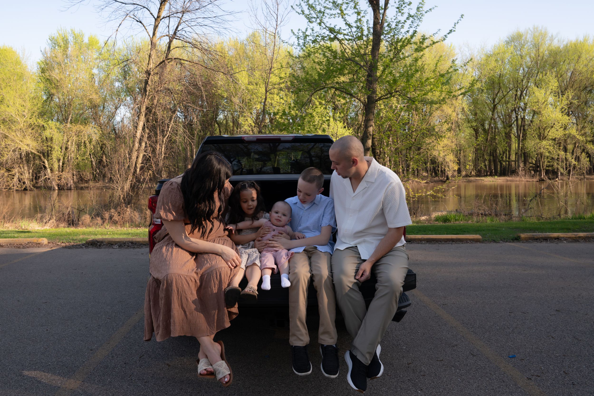 Family of five sitting on a truck tailgate in a parking lot surrounded by green trees near a pond.