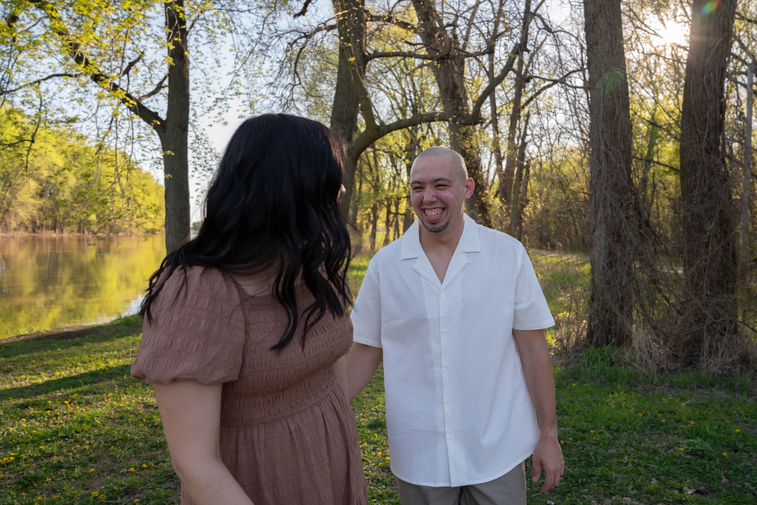 Couple smiling outdoors near a sunlit lake surrounded by trees in a lush green park setting.