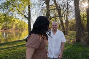 A smiling bald man in a white shirt faces a woman with dark hair in a brown dress by a sunlit river.