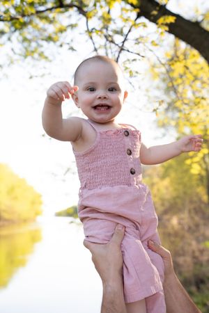 Happy baby in pink romper being held up outdoors near a sunlit river, arms outstretched and smiling joyfully.
