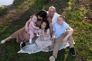 Happy family of five sitting on a blanket outdoors on green grass, parents with three young children smiling together.
