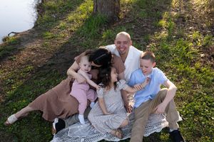 Happy family of five sitting on a blanket by a riverside, laughing and cuddling in warm golden sunlight.